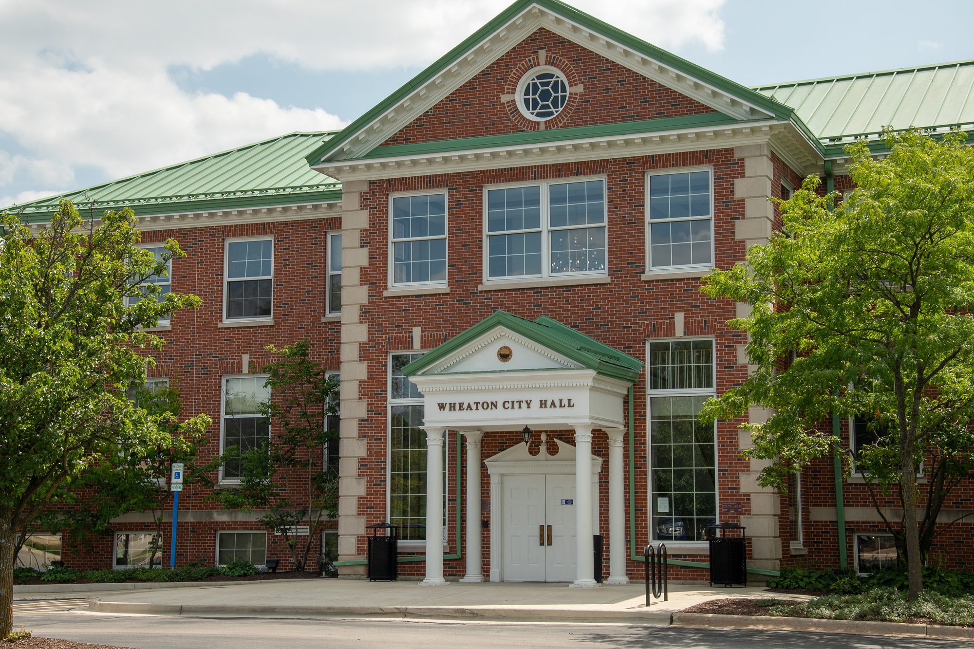 Two-story red brick city hall with white columned entrance and green metal roof.