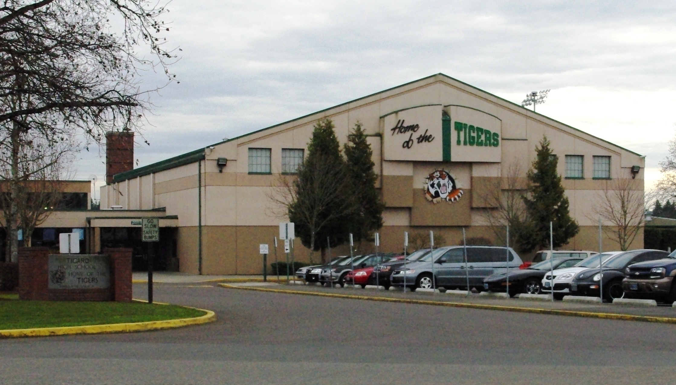 School building exterior with 'Home of the Tigers' sign, parking lot, and evergreen trees.