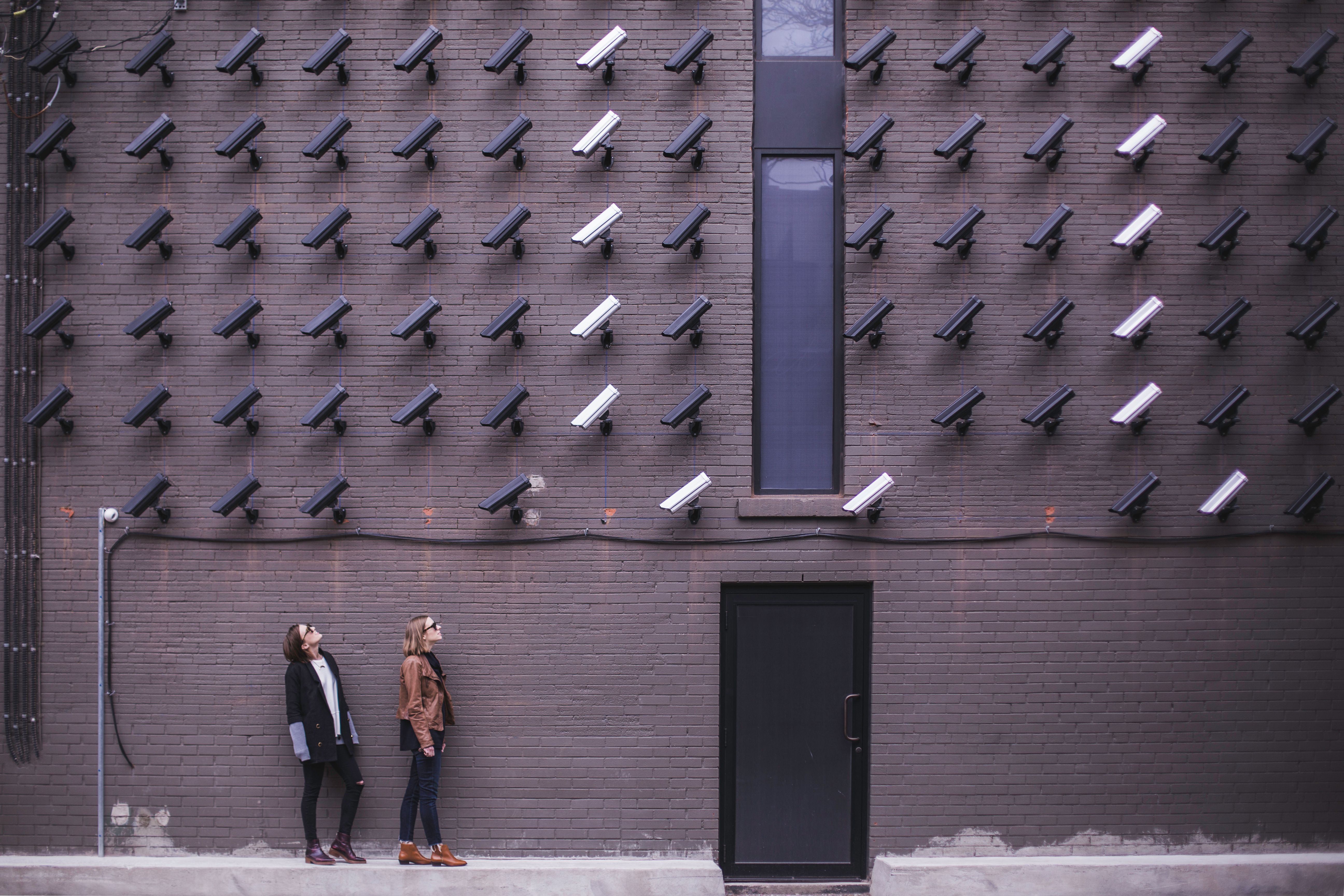 Two people look up at dozens of surveillance cameras mounted on a brick wall.