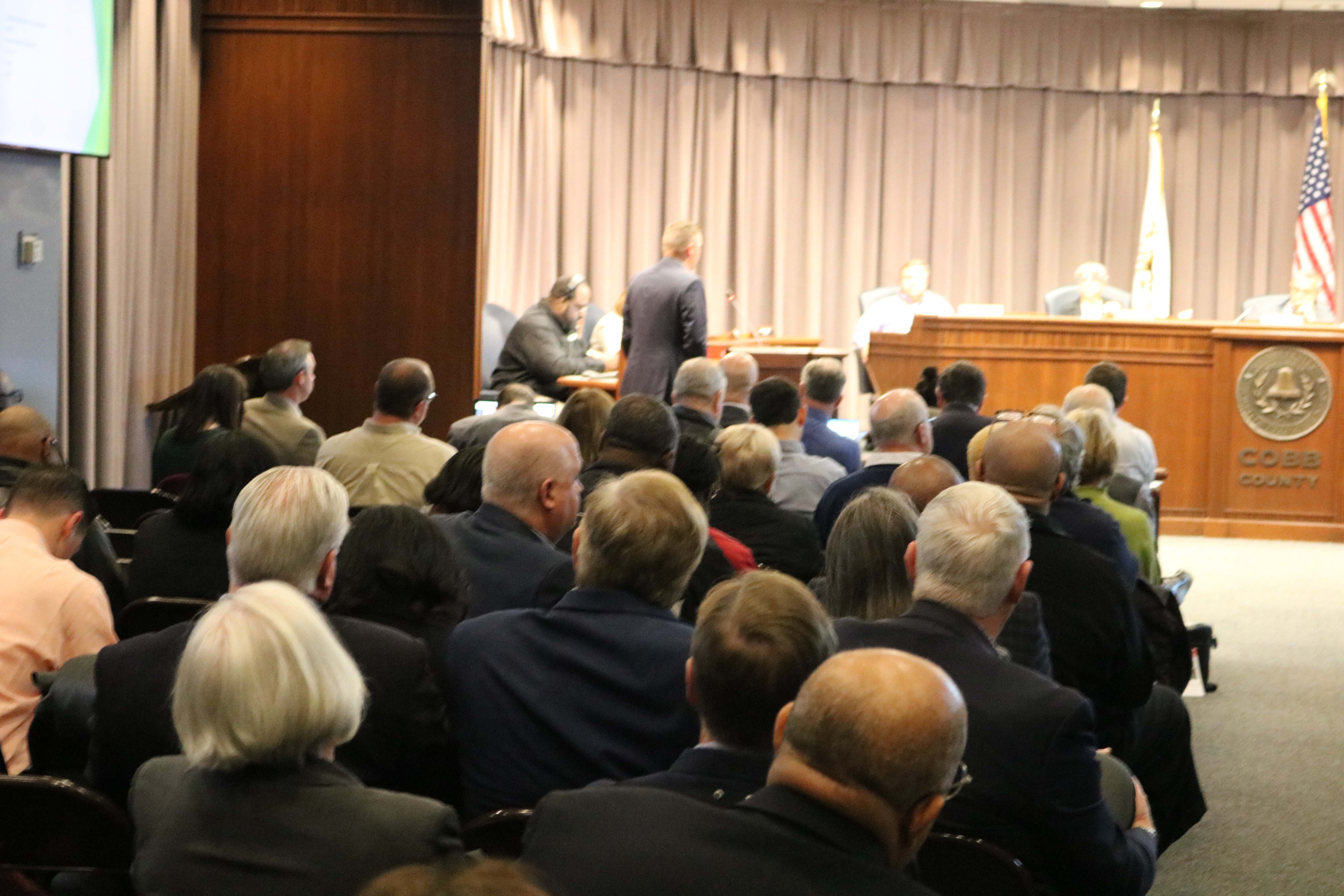 Audience seated in rows facing a raised government dais with an American flag and county seal.