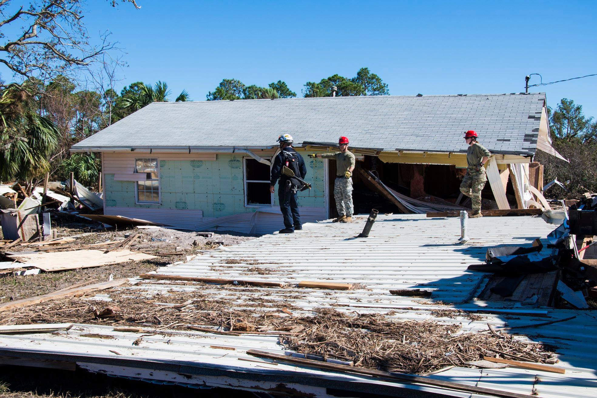 Three uniformed rescuers in red helmets stand outside a heavily storm-damaged house.