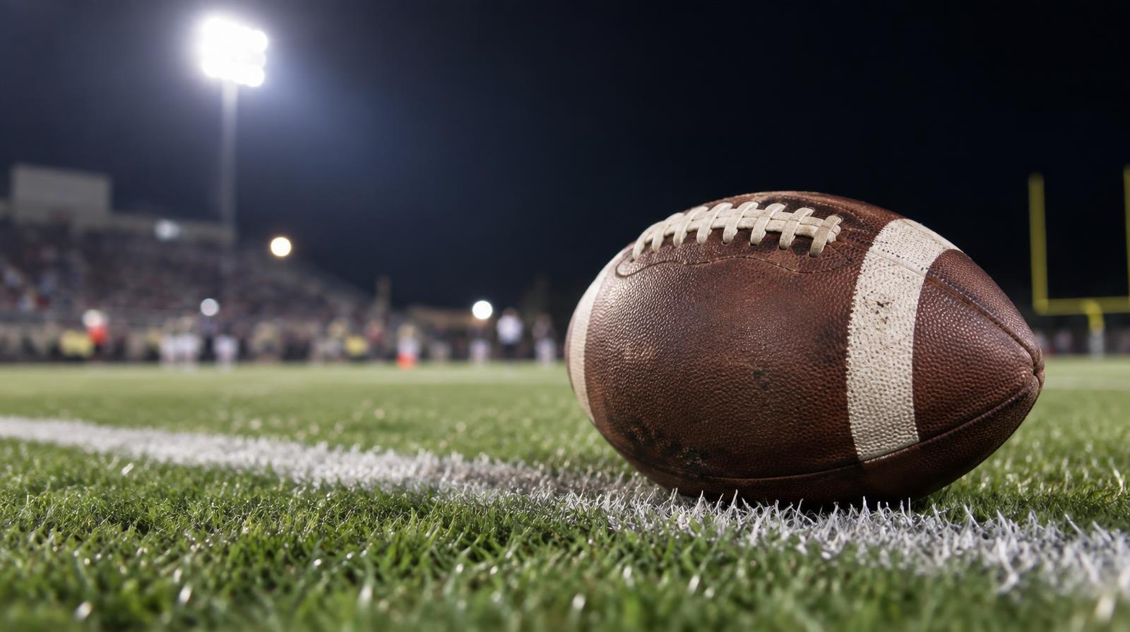 An American football on green grass near a white yard line under bright lights.