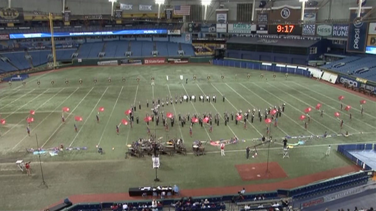 Marching band with red color guard flags in a circular formation on a stadium field, viewed from above.