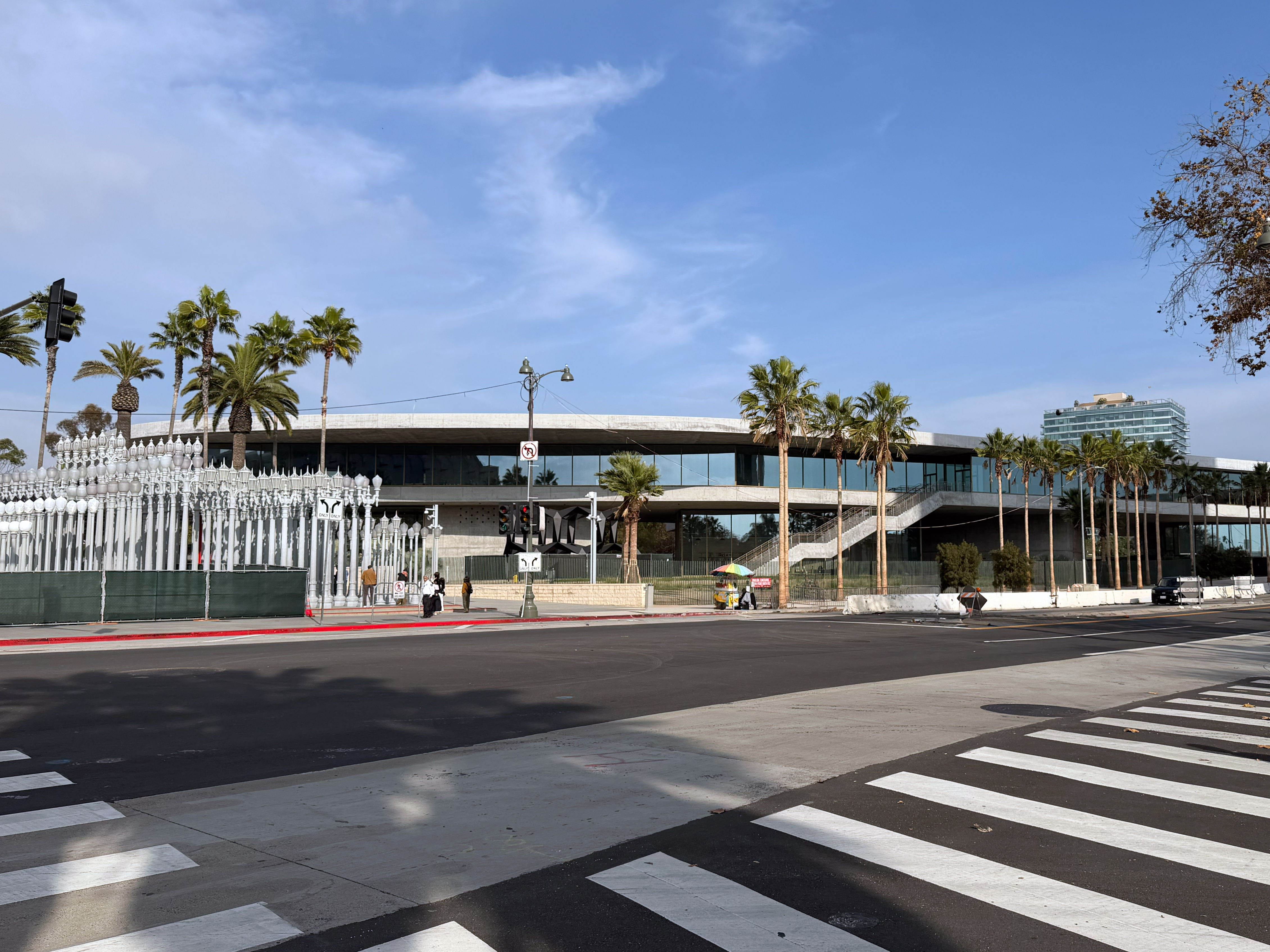 Modern glass-and-steel museum building with palm trees and lamp post sculptures at the entrance.