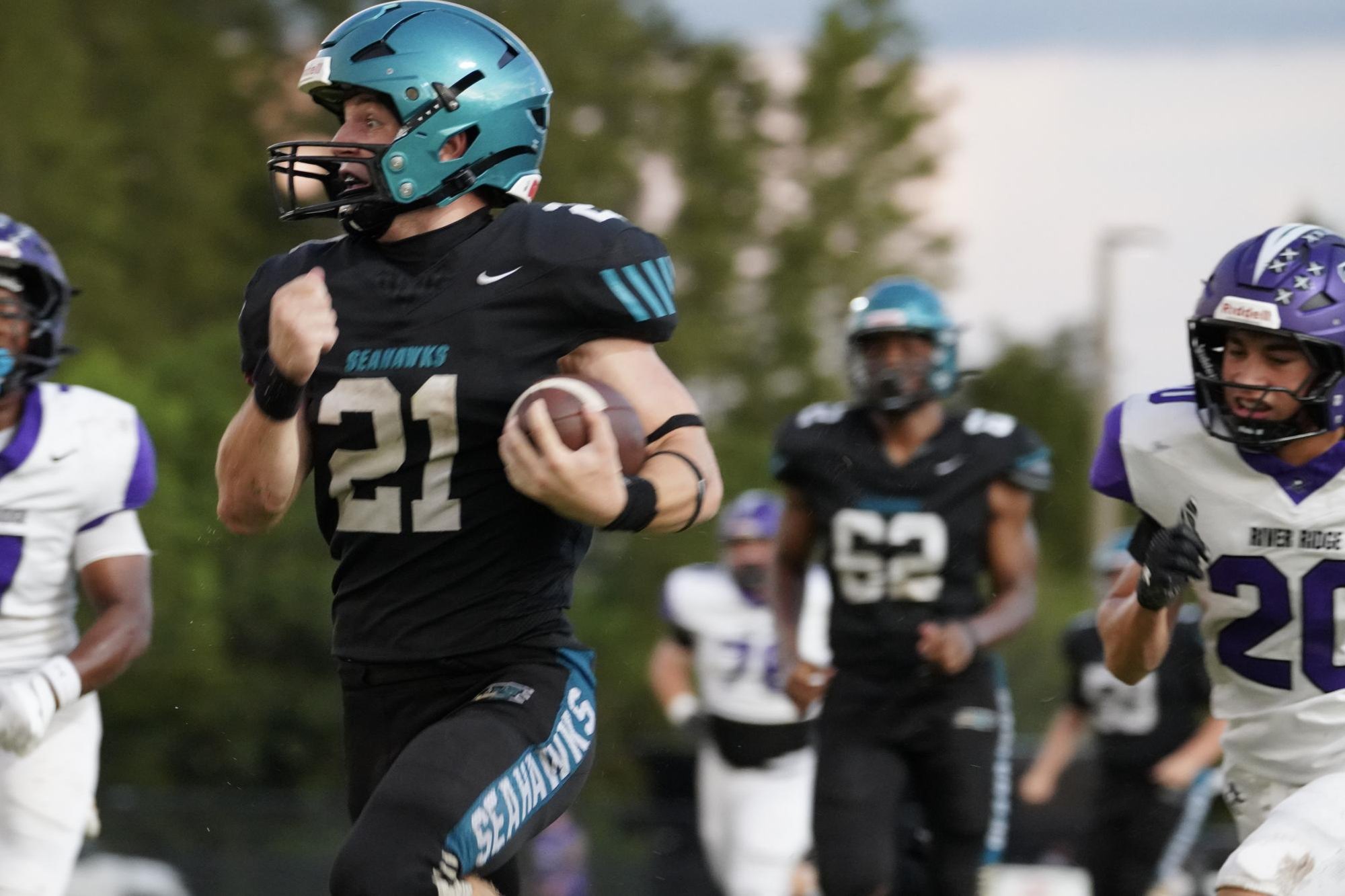 Football player in black Seahawks jersey number 21 runs with the ball pursued by River Ridge defenders.