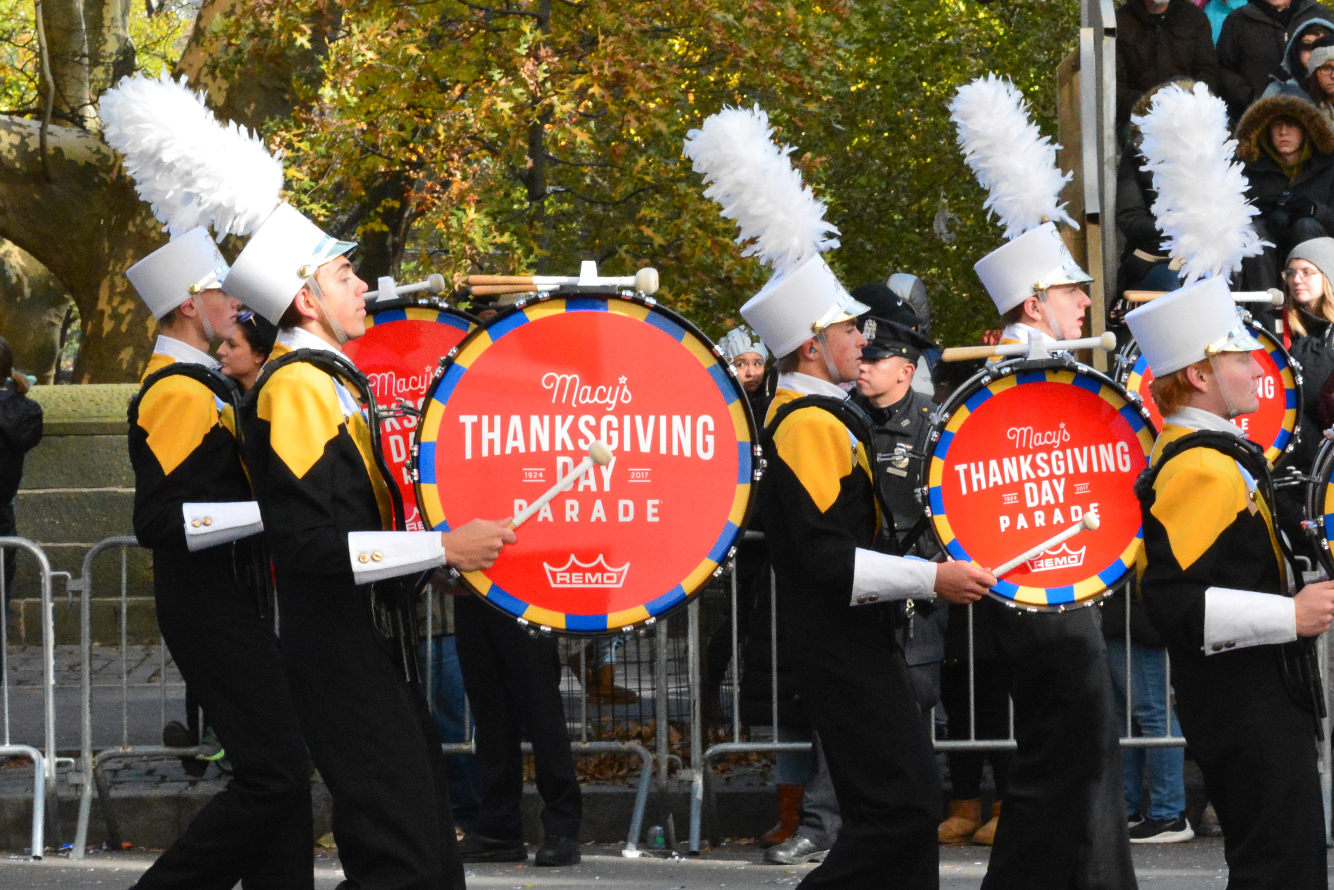 Marching band members in black-and-gold uniforms carry large Macy's parade bass drums.