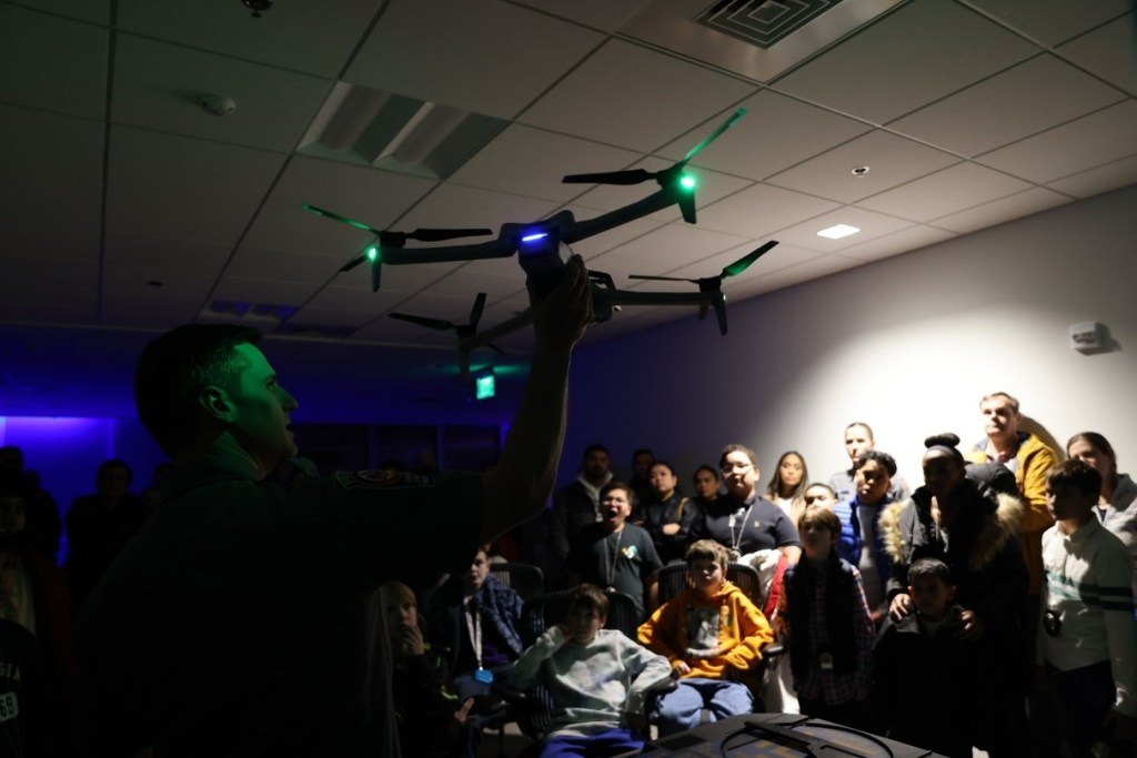 A man holds a large drone with green lights while a crowd of adults and children watches indoors.