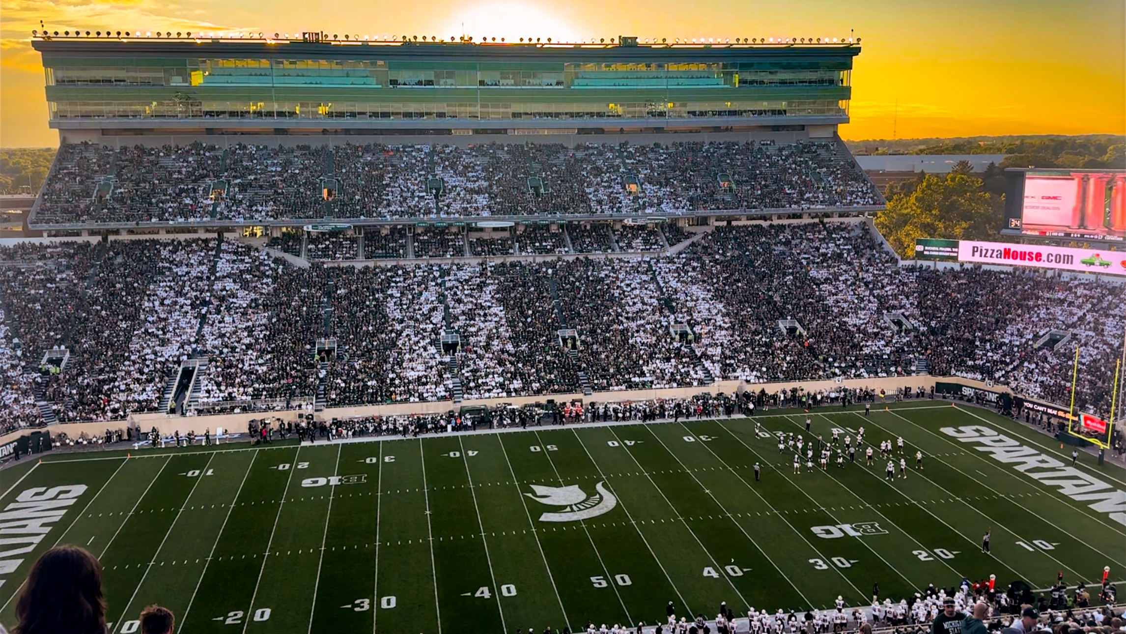 Packed football stadium at sunset with fans in white and a green playing field.