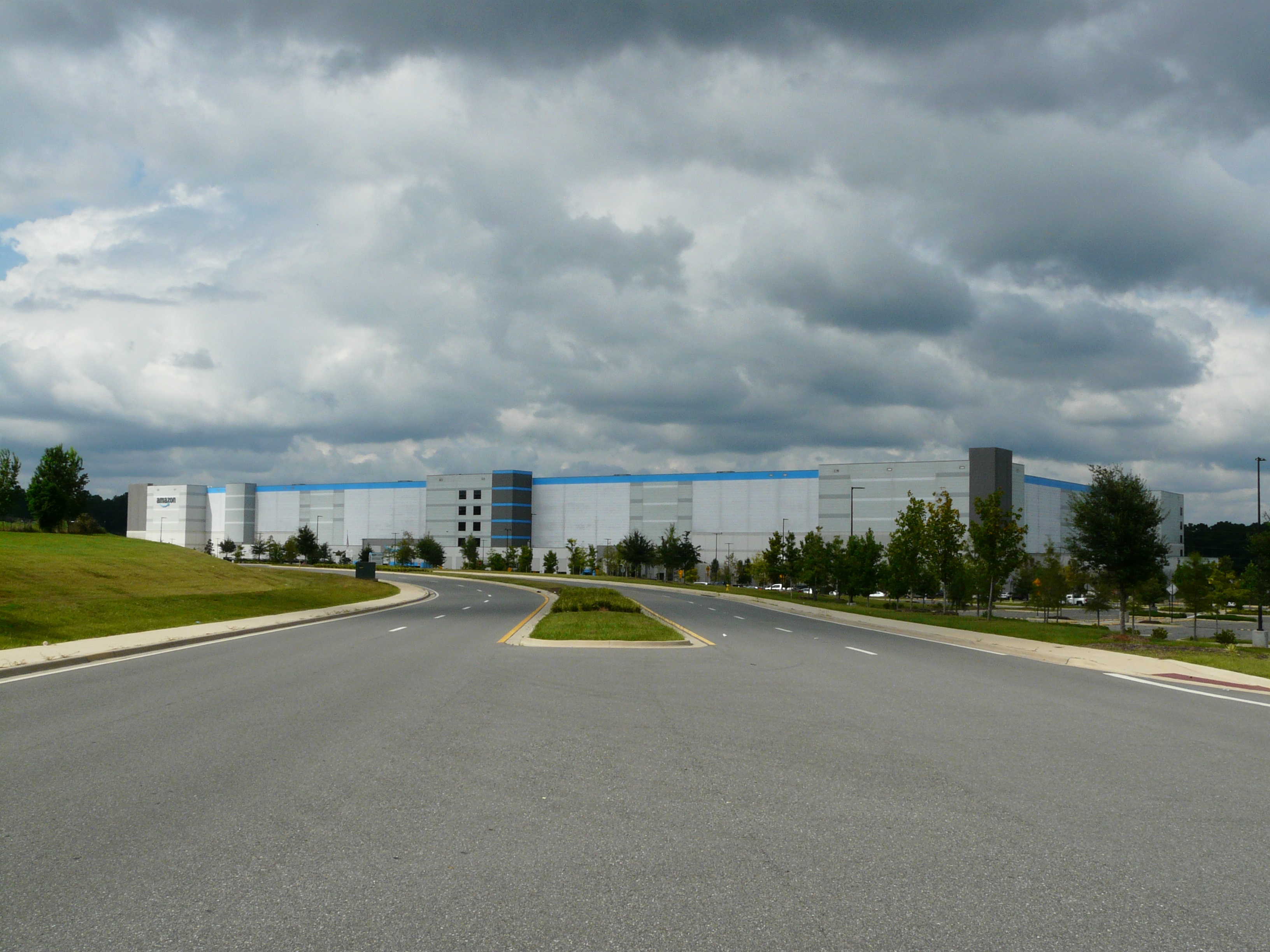 Large blue and white warehouse building viewed from a wide multi-lane road with landscaped medians under overcast skies.