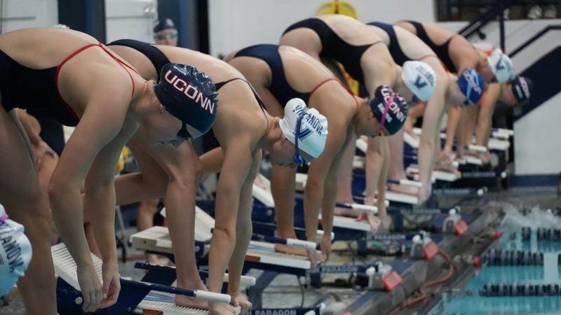 Female swimmers crouched on starting blocks at an indoor pool, wearing swim caps and goggles.