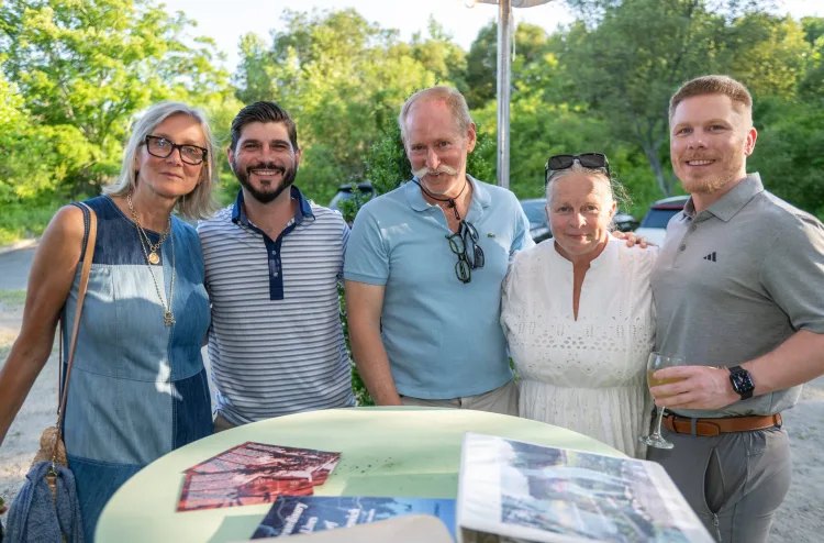 Five adults smiling together at an outdoor summer gathering with greenery behind them.