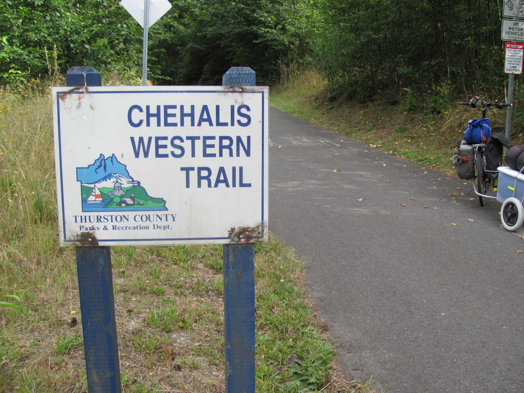 Trail entrance sign on blue posts with a paved path and parked bicycle behind it.