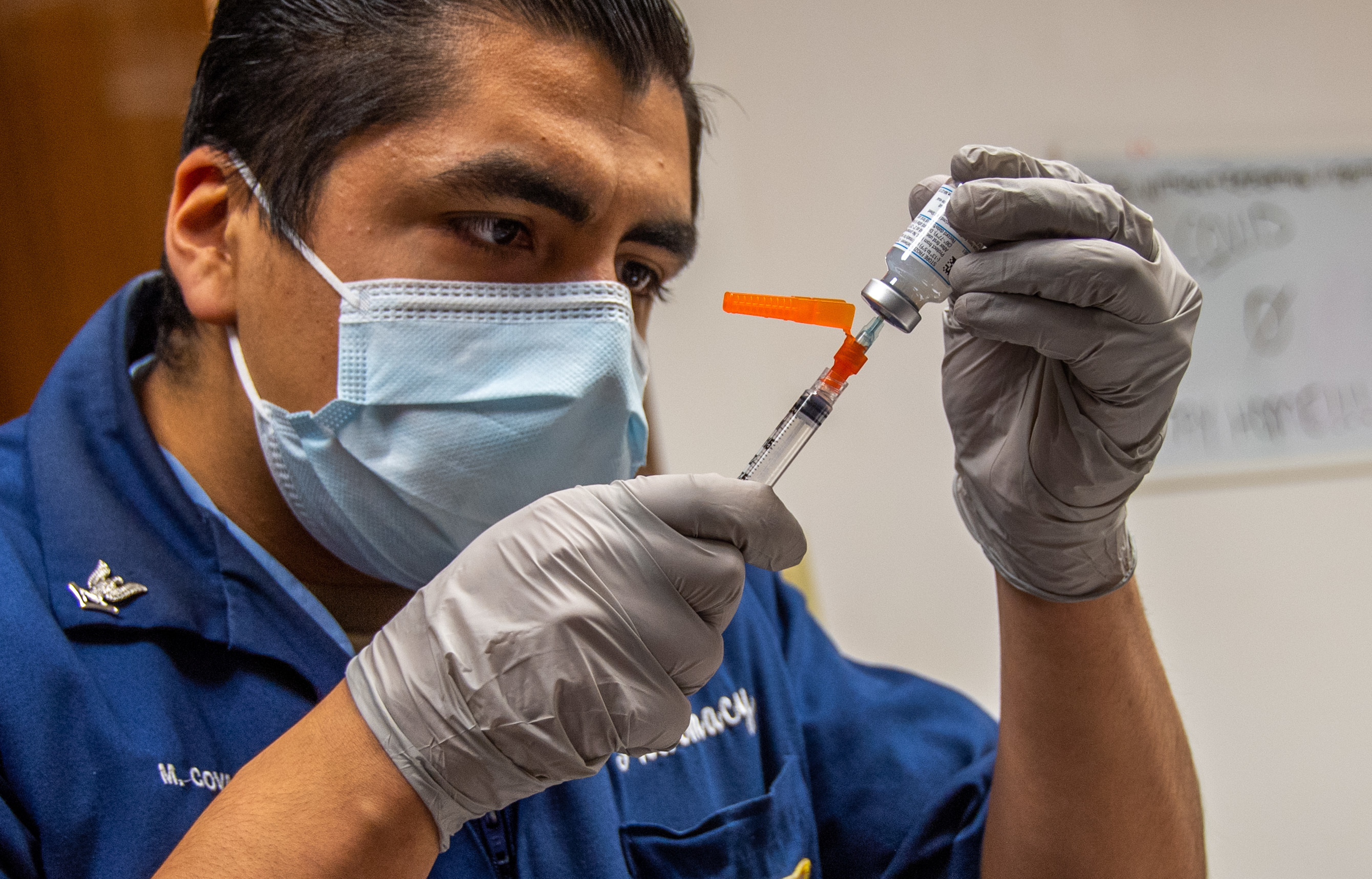 Masked healthcare worker in blue uniform drawing vaccine into a syringe from a vial.