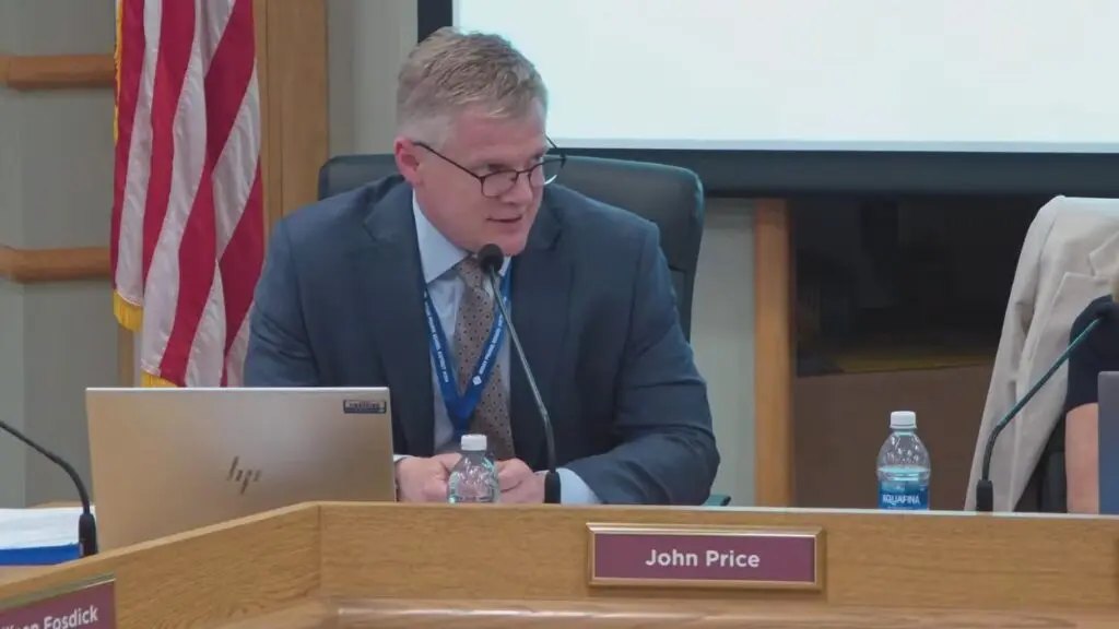 Man in dark suit and glasses seated at a board meeting dais behind a nameplate reading John Price.