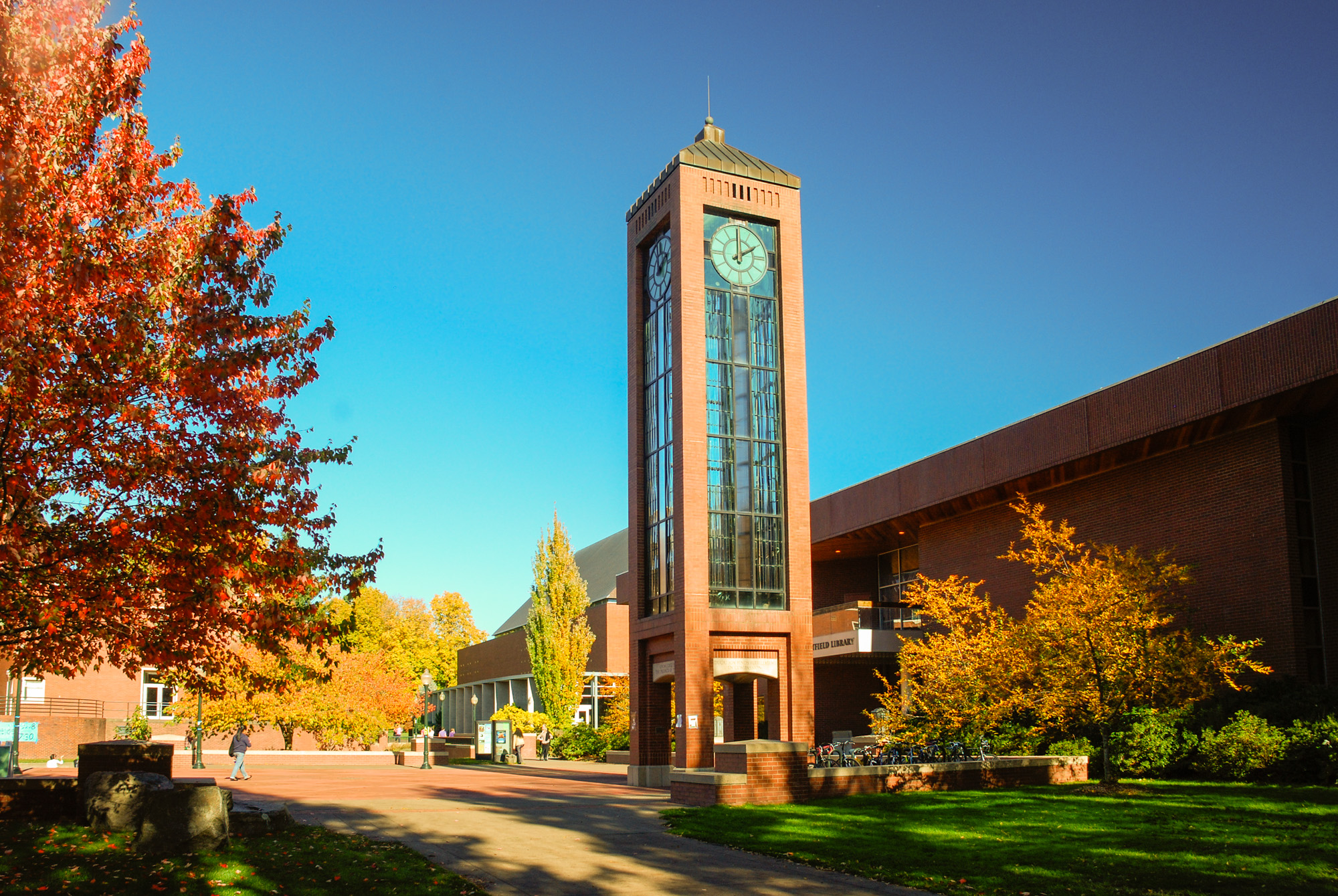 Brick and glass clock tower on a university campus surrounded by autumn trees.