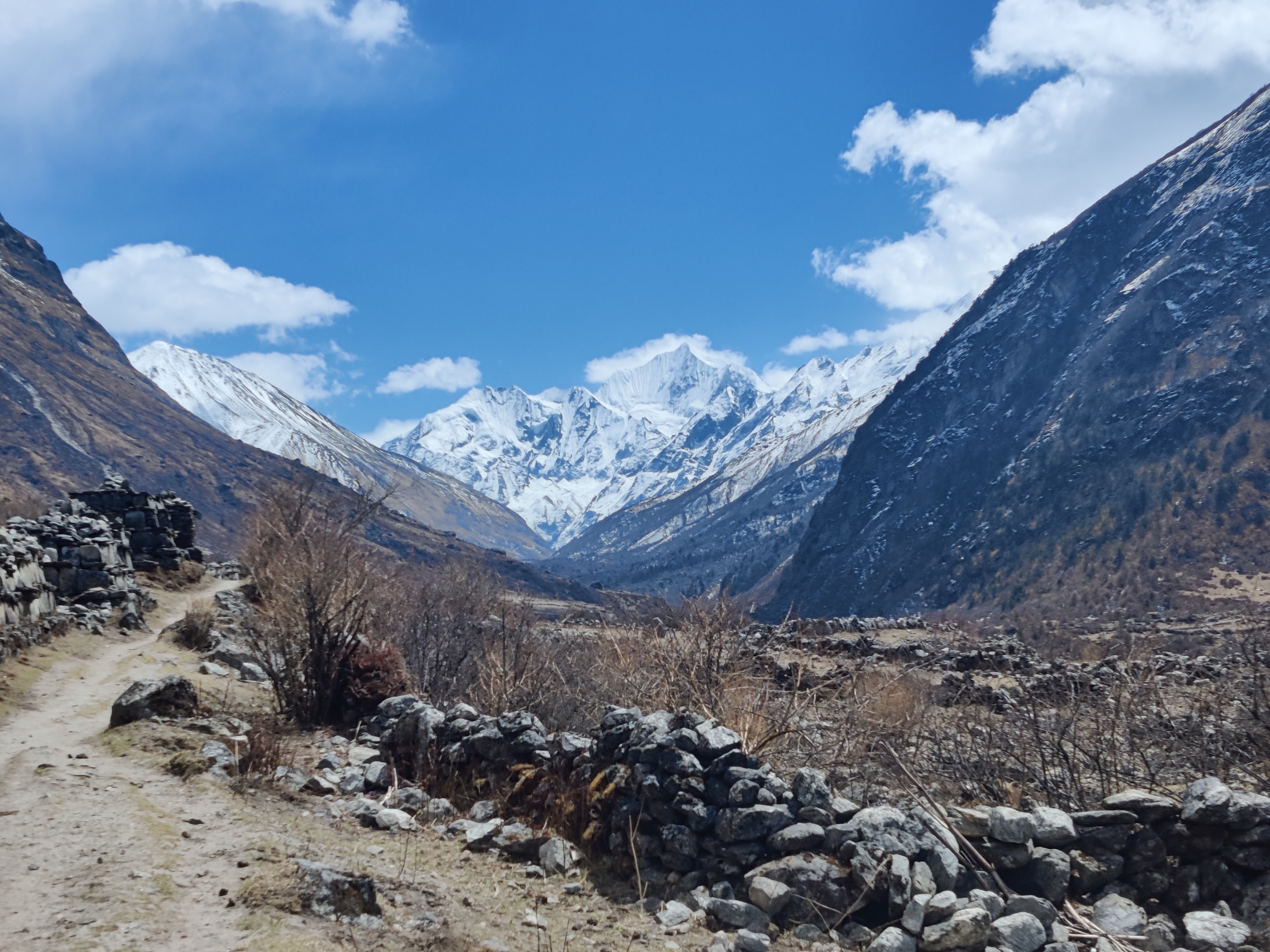 A rocky mountain trail with a stone wall and snow-capped peaks in the distance.