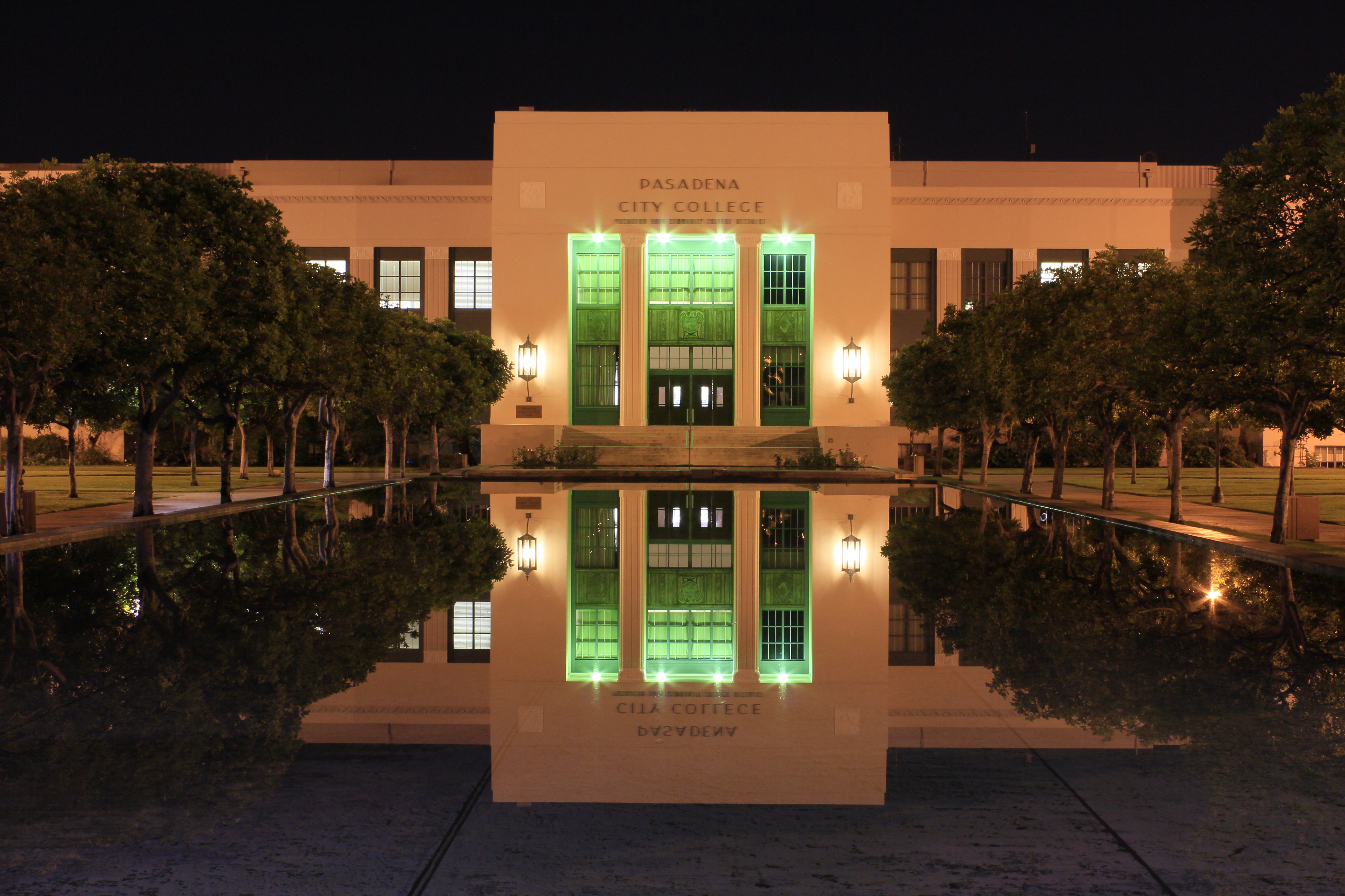 Illuminated campus building with green-lit columns reflected in a pool at night.