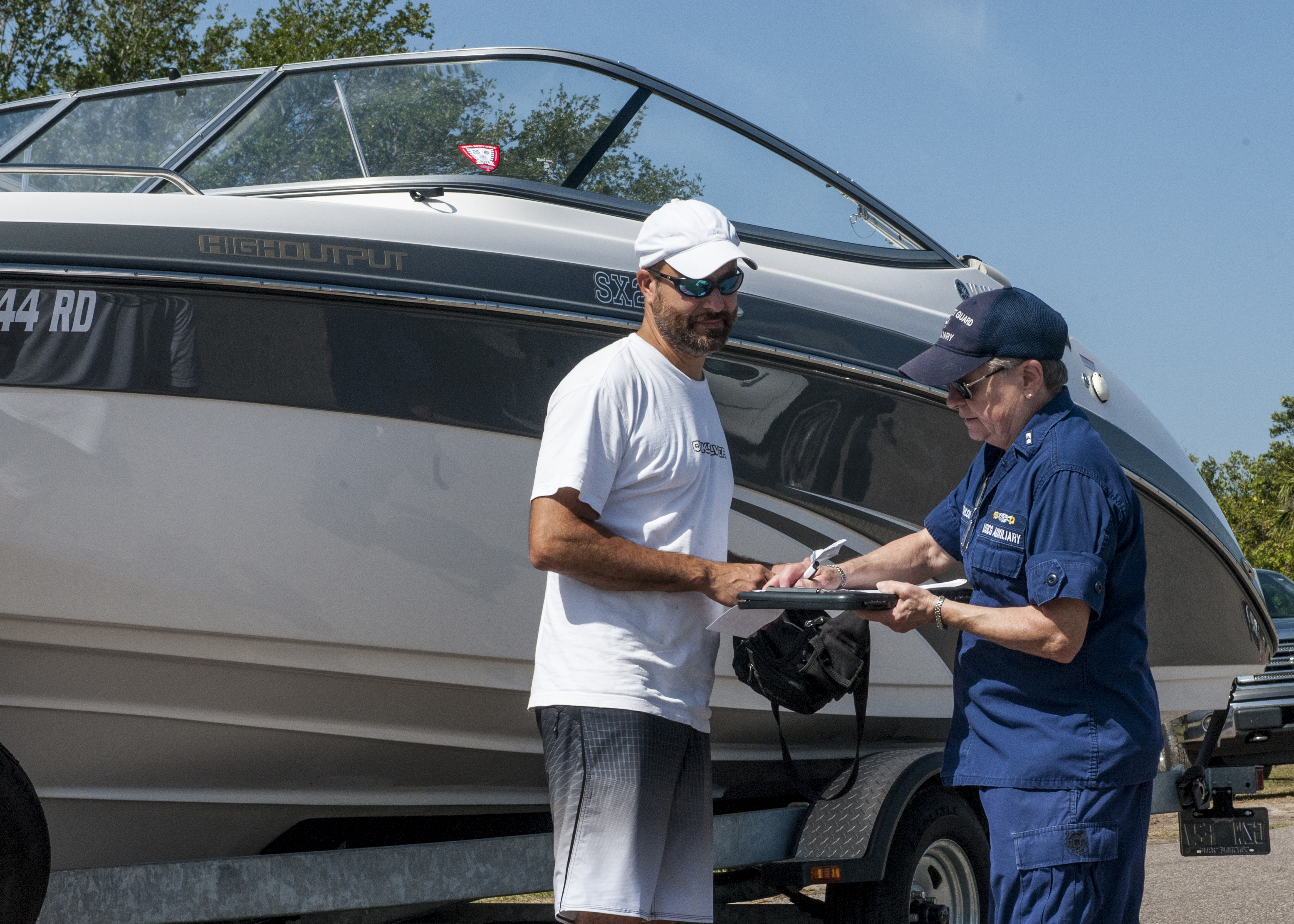 Coast Guard Auxiliary member in uniform holds clipboard while speaking with a boat owner outdoors.