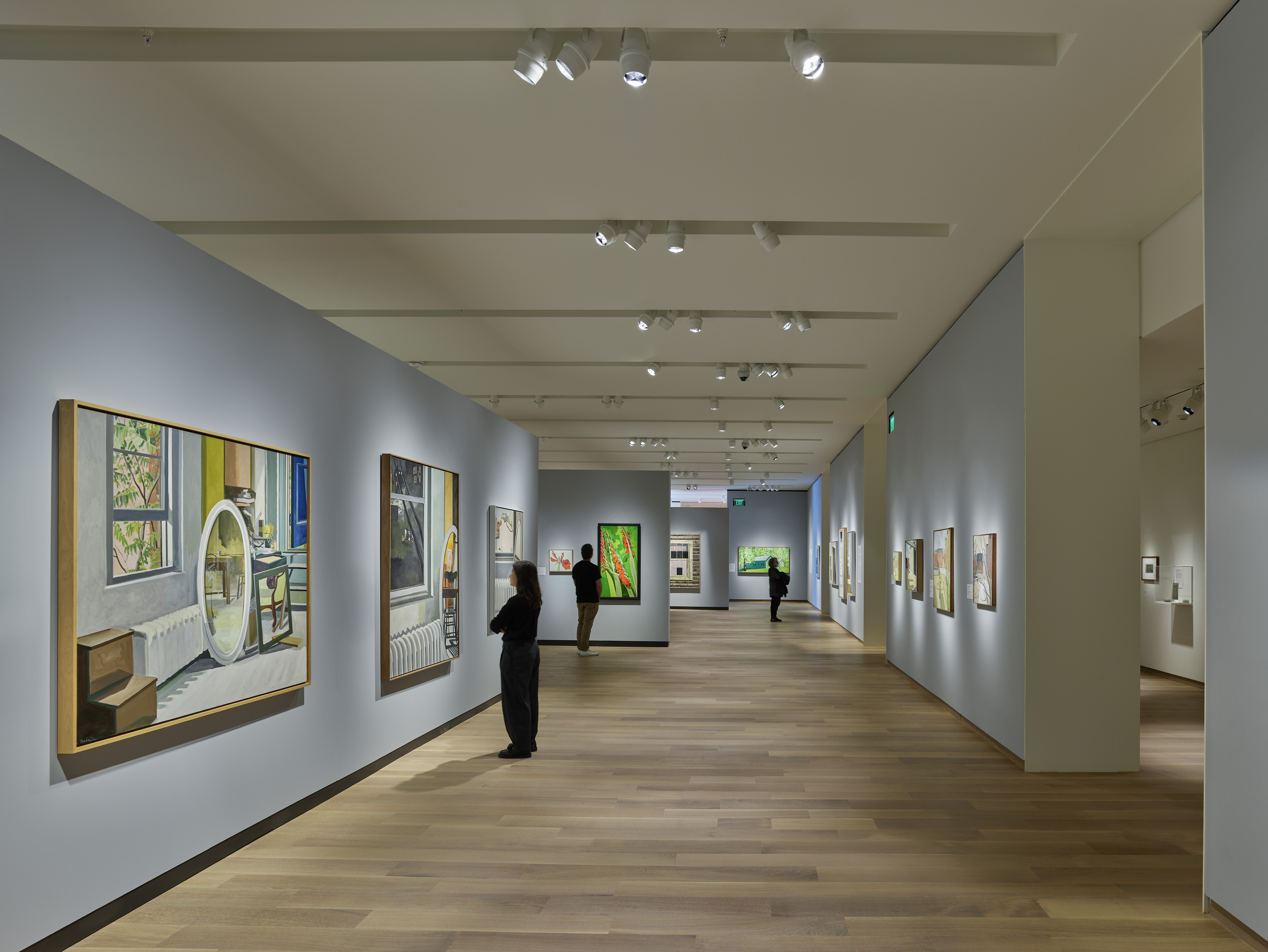 A museum gallery corridor with grey walls, track lighting, and paintings on both sides.