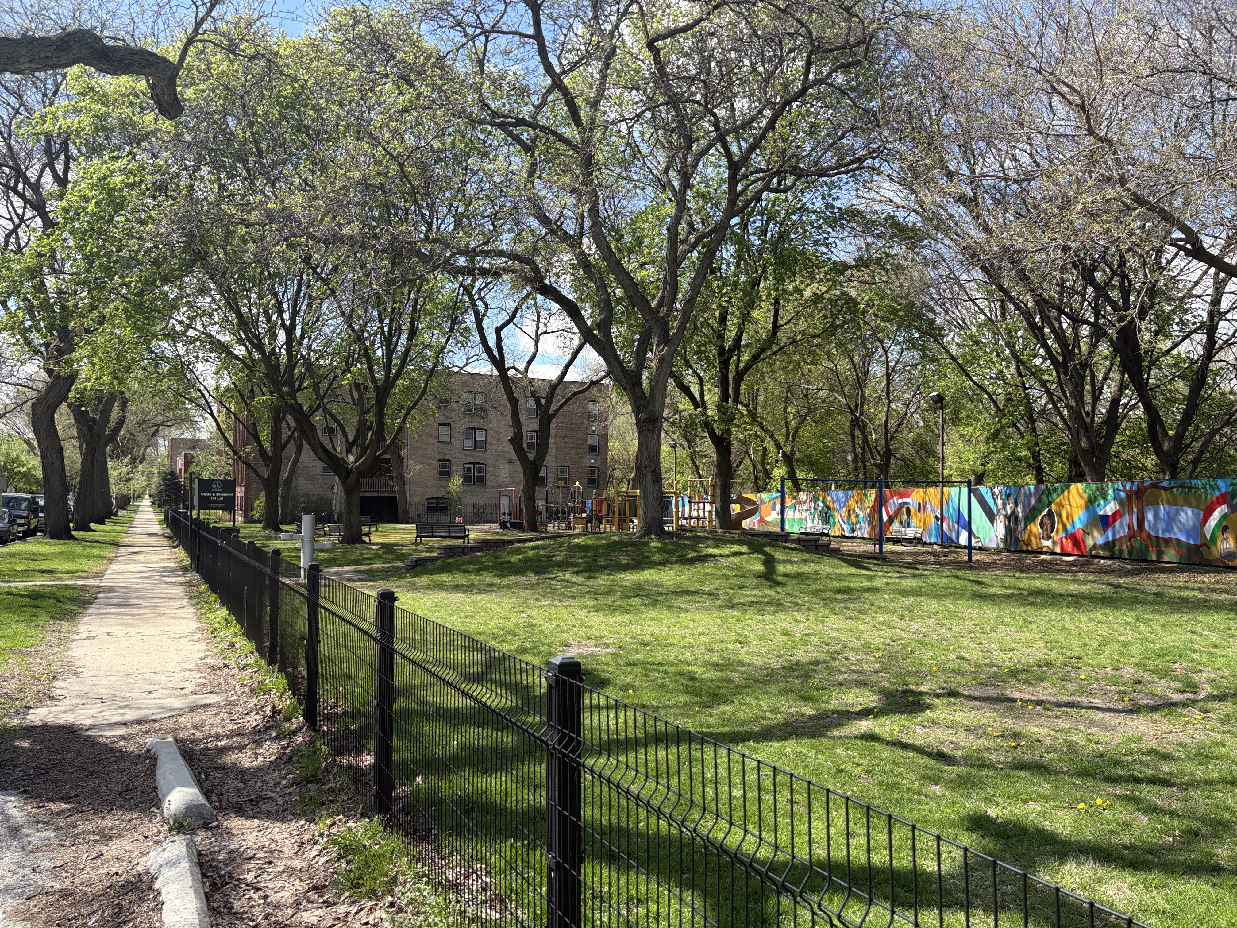 Grassy park with mature trees, black metal fence along a paved path, and a colorful mural on a background fence.