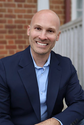 Robert Jones pictured sitting on a porch, smiling, and wearing a dark blue (navy) blazer with a light blue, button-down shirt.