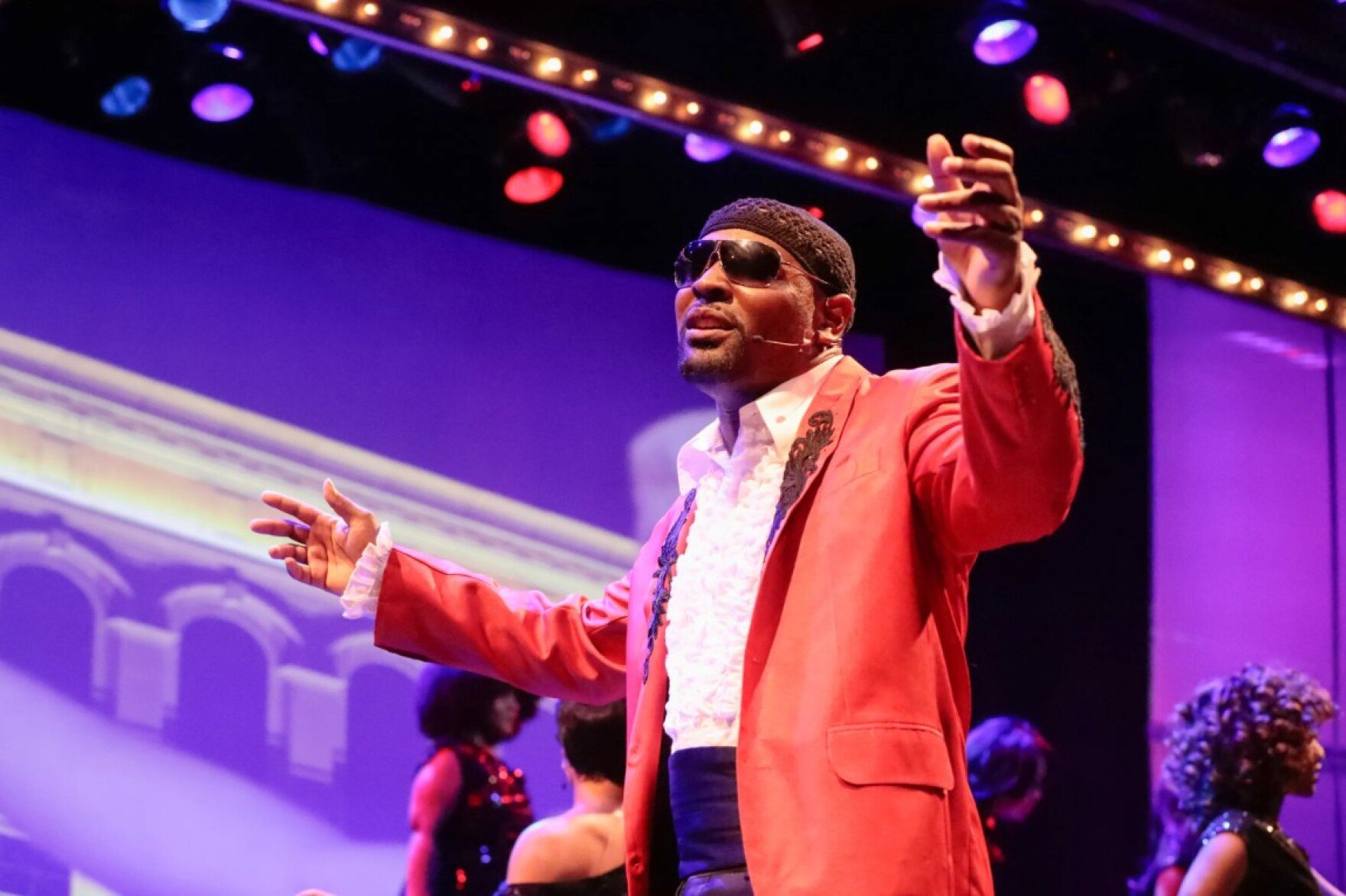 A performer in sunglasses and a peach suit jacket sings on stage with arms outstretched under colorful lights.
