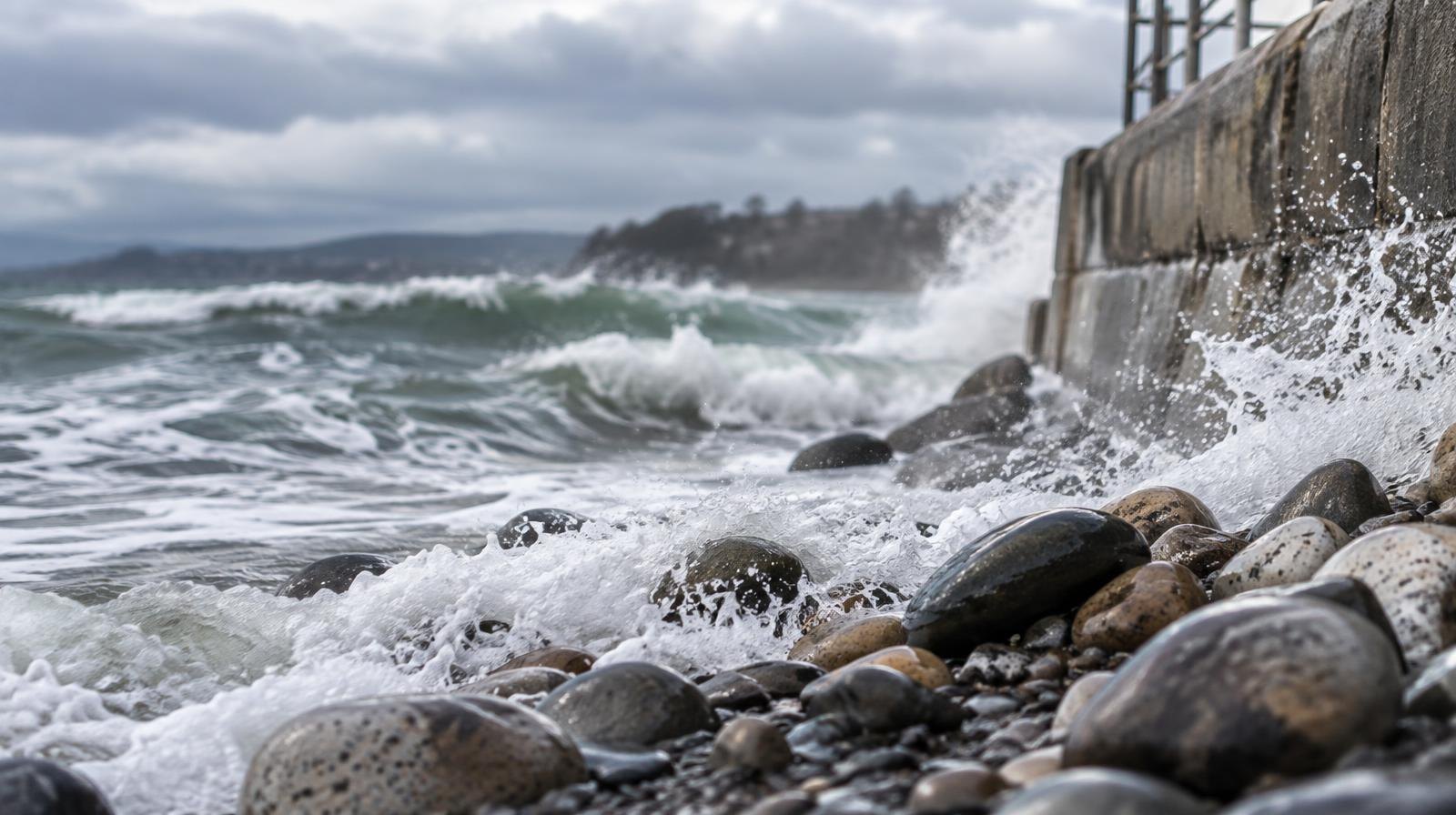 Rocky pebble-covered beach with rough surf washing over cobblestones at a concrete seawall.