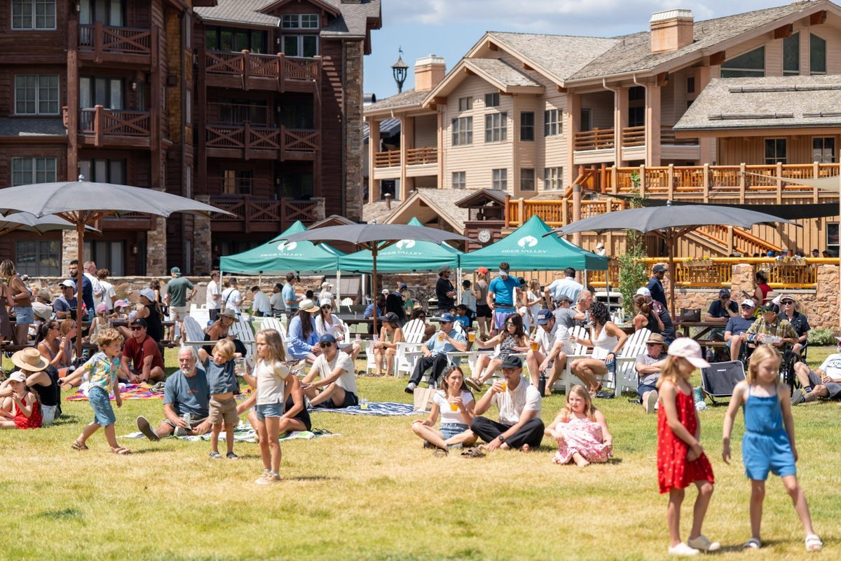 Crowd of adults and children on a sunny lawn at a mountain resort with green vendor tents.