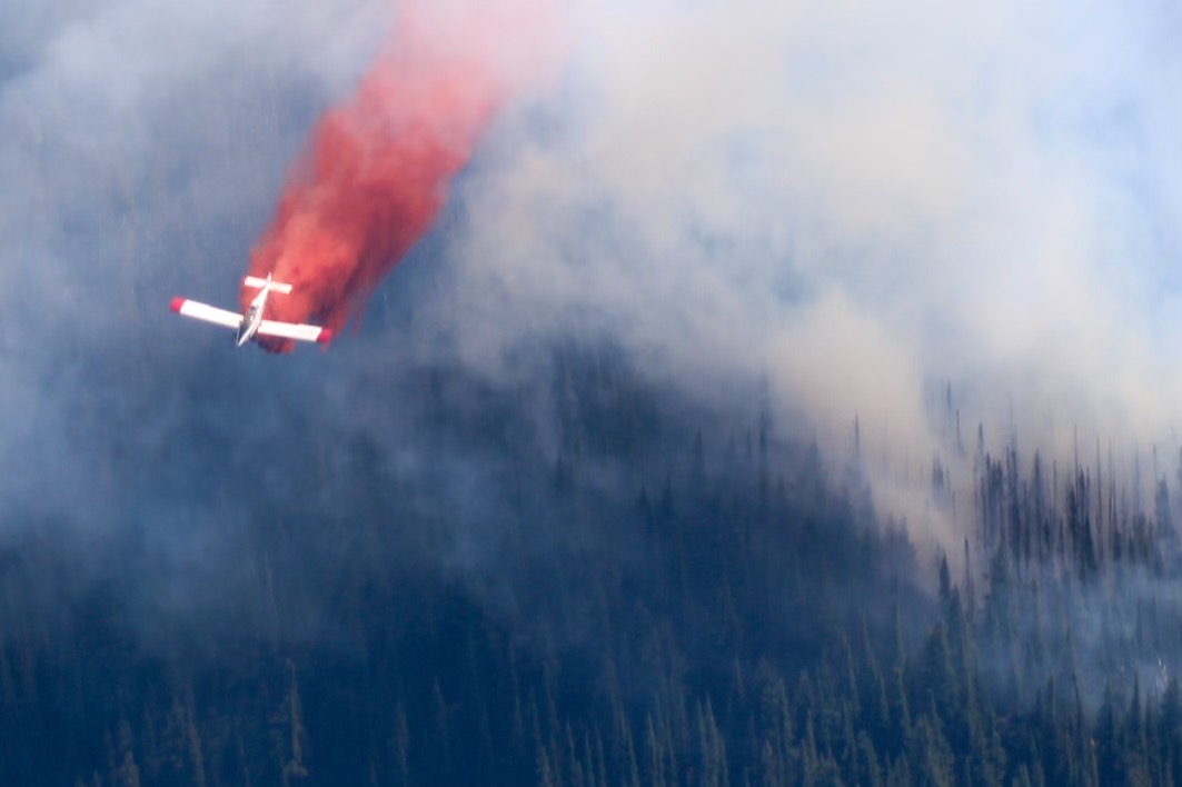 Small fixed-wing aircraft releasing red fire retardant over a smoky, forested hillside.