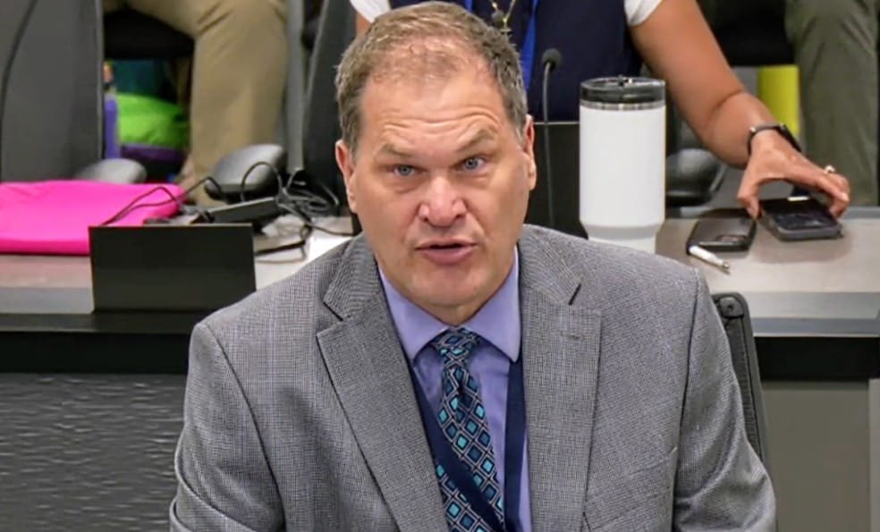 A man in a gray suit and patterned tie speaks at a government meeting.