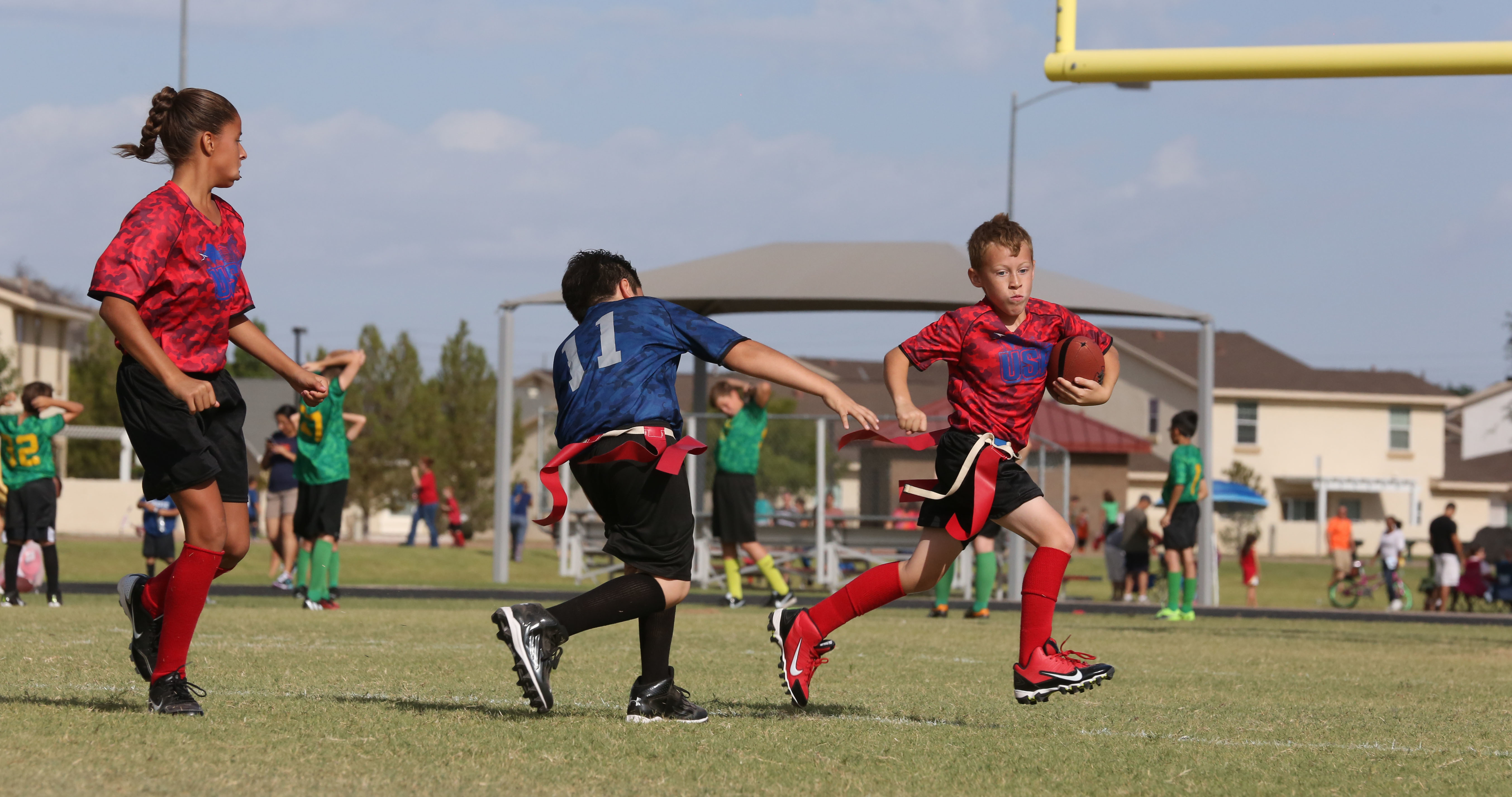 A child in a red jersey carries a football while a defender reaches for his flag belt on a grass field.