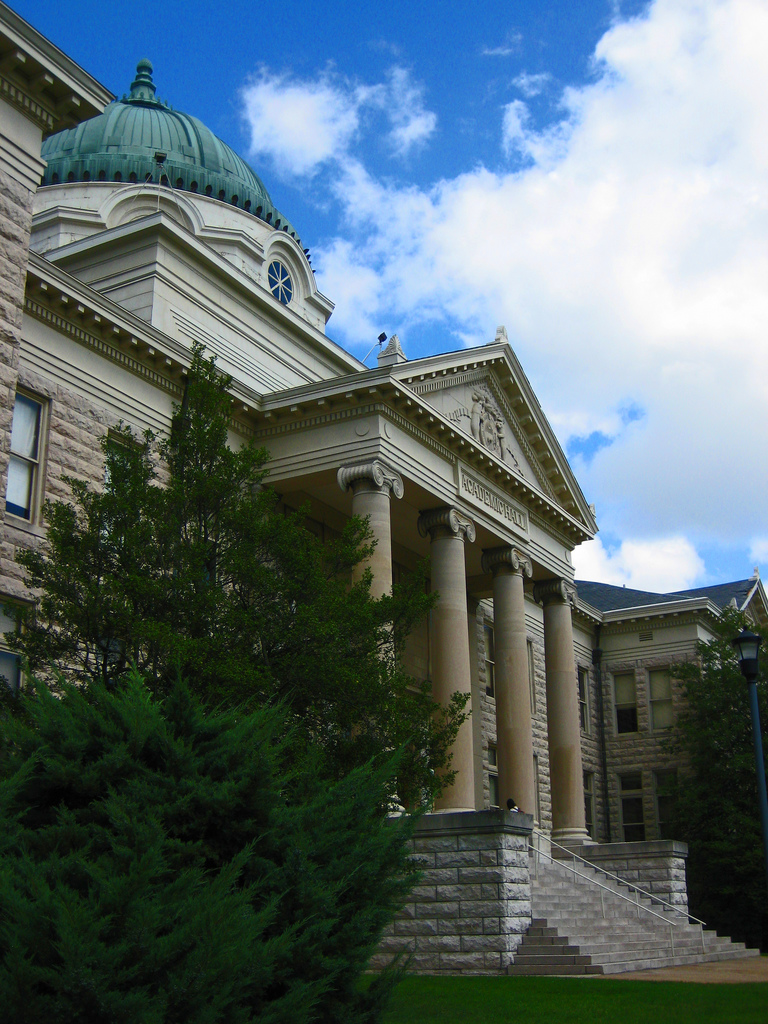 Stone university building with a green copper dome and columned entrance, flanked by trees.
