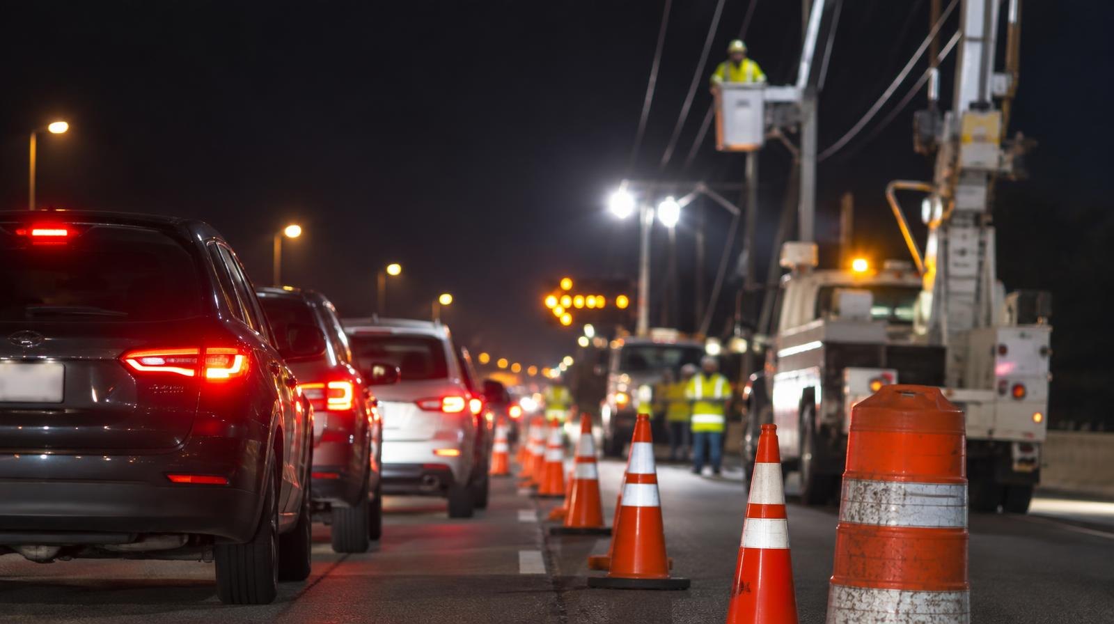 Orange traffic cones along a dark highway at night with work crew lighting.