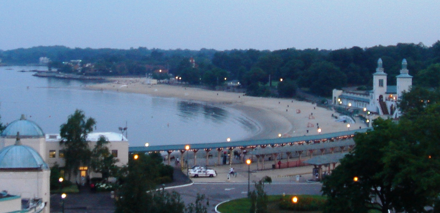Curved sandy beach and boardwalk at dusk with illuminated street lights along the waterfront.