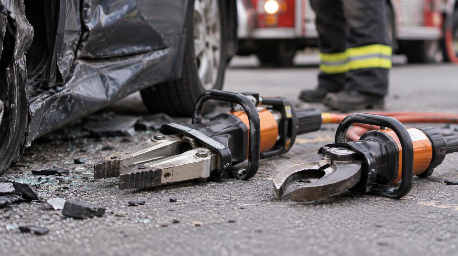 Hydraulic spreader and cutter tools beside a crumpled car door on pavement.