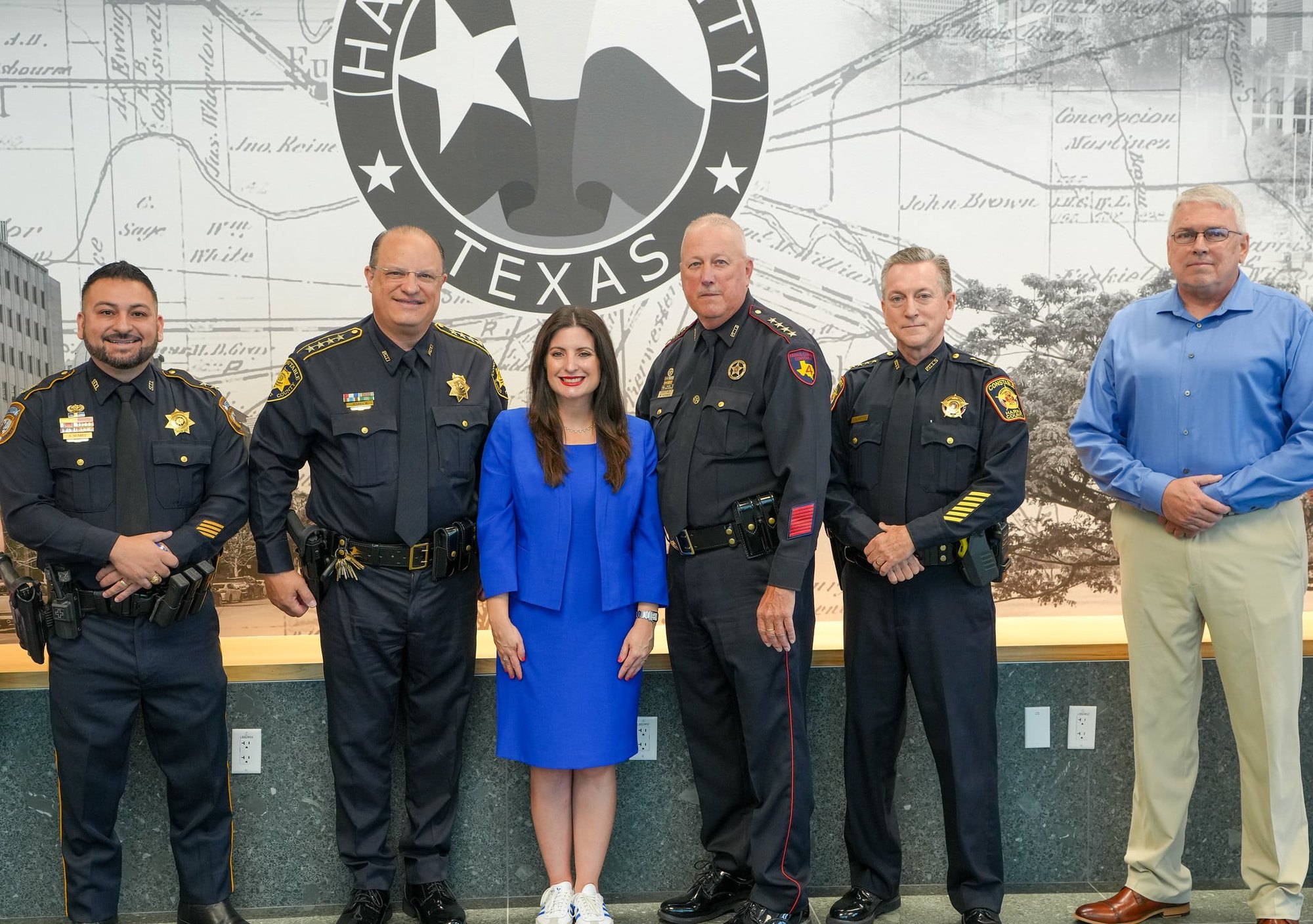 Four uniformed law enforcement officers and two civilians pose in front of a Harris County seal mural.