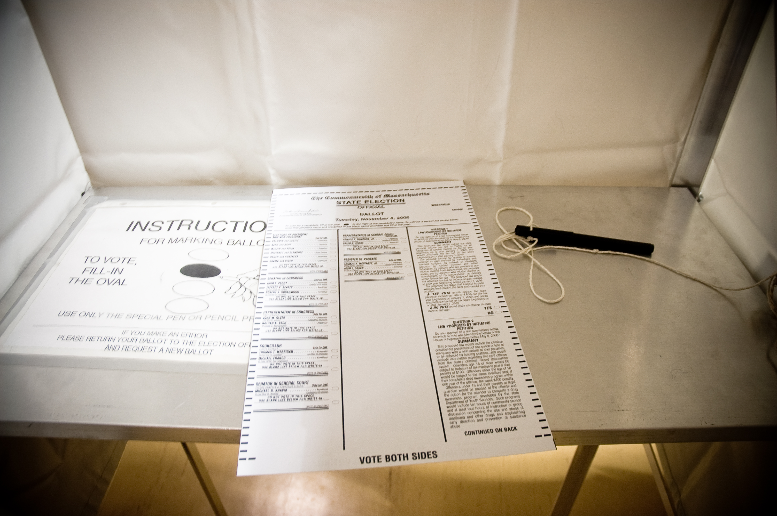 Inside a voting booth, a printed ballot and pen rest on a metal shelf.
