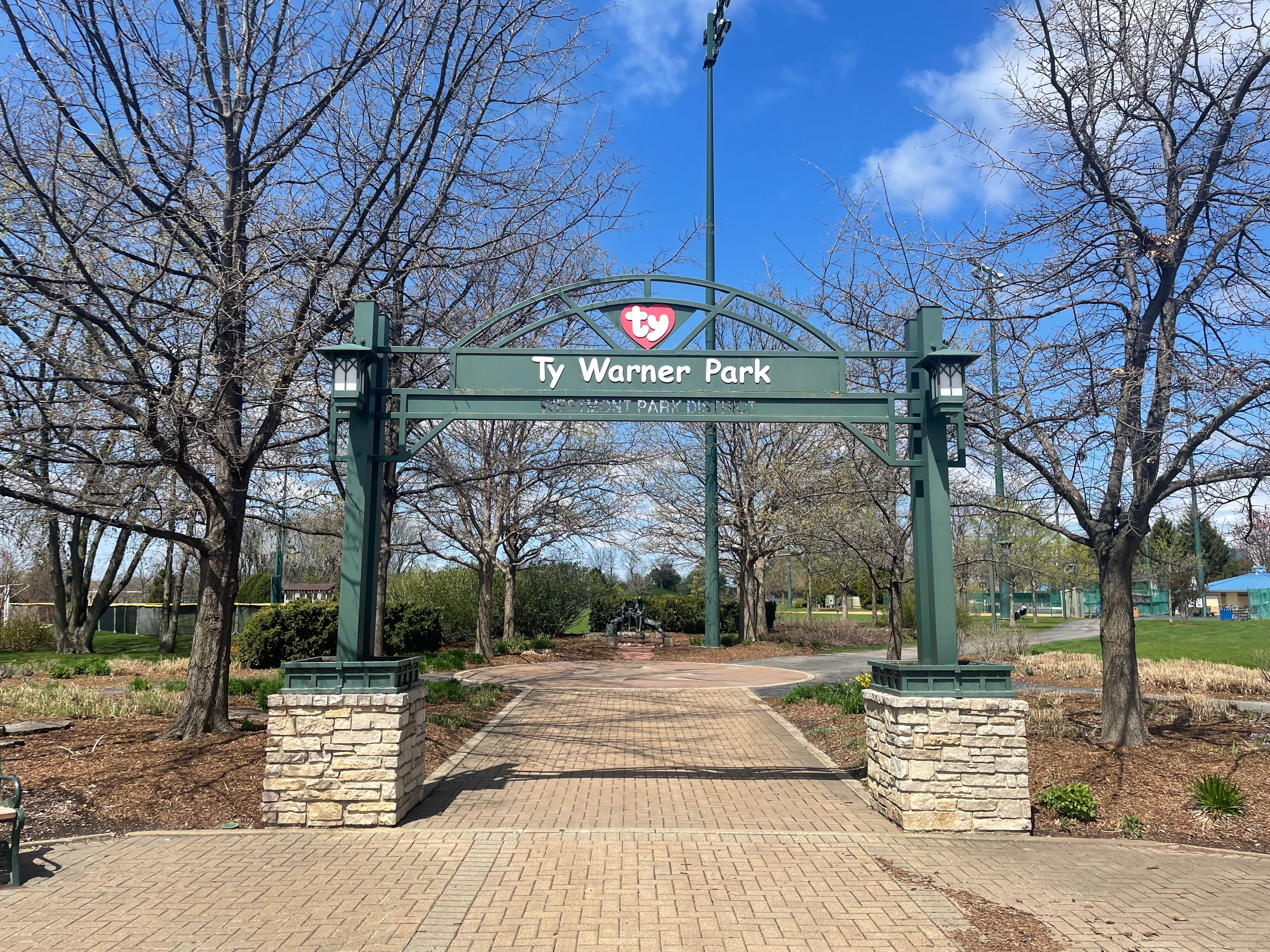 Green metal arch reading Ty Warner Park with stone pillars and a brick walkway leading into the park.