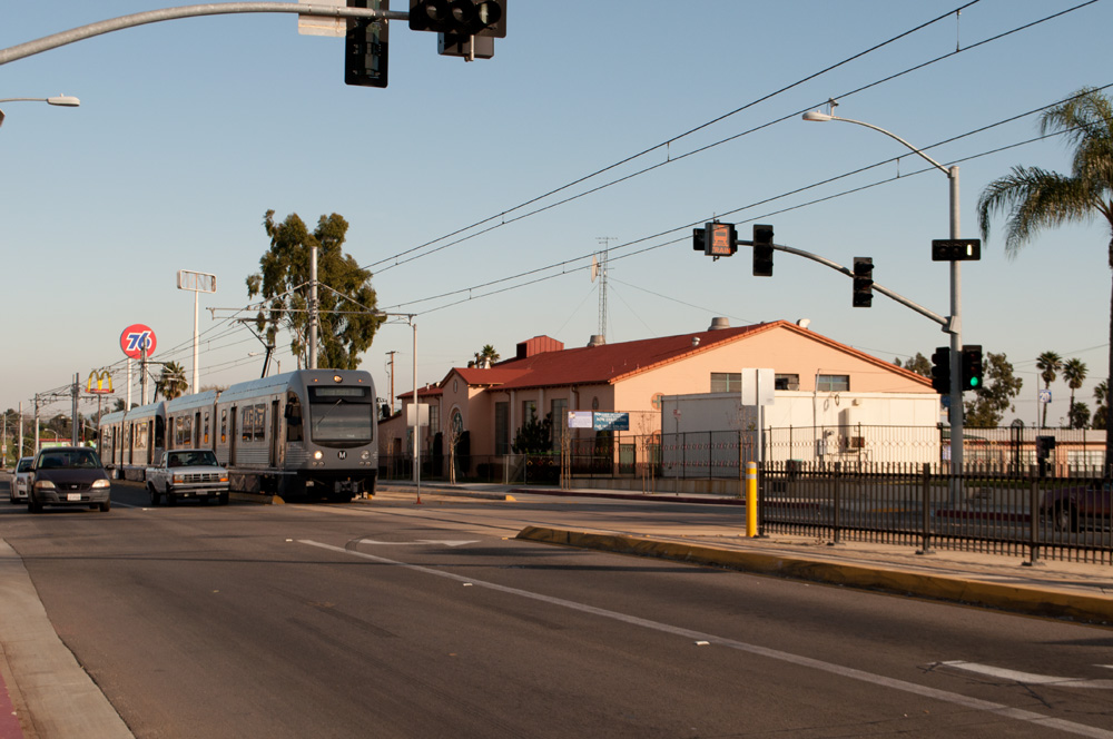 A Metro light rail train travels along a street-level track at an urban intersection.