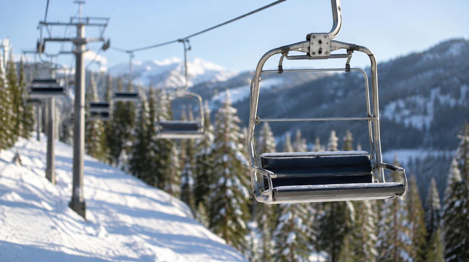 Empty ski lift chairs suspended above a snowy slope lined with pine trees.