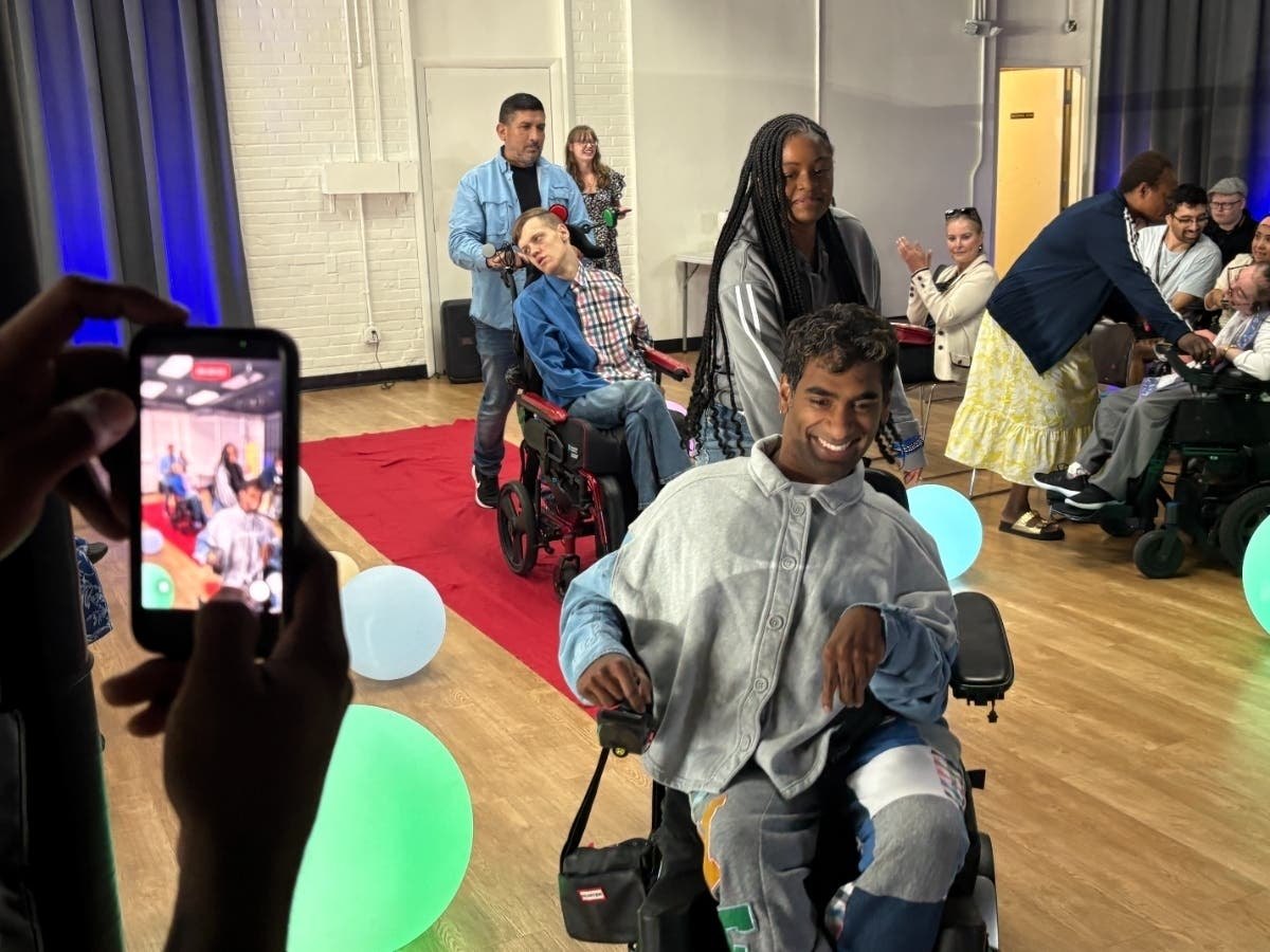 People in power wheelchairs roll along a red carpet runway inside an indoor venue with glowing balloons.