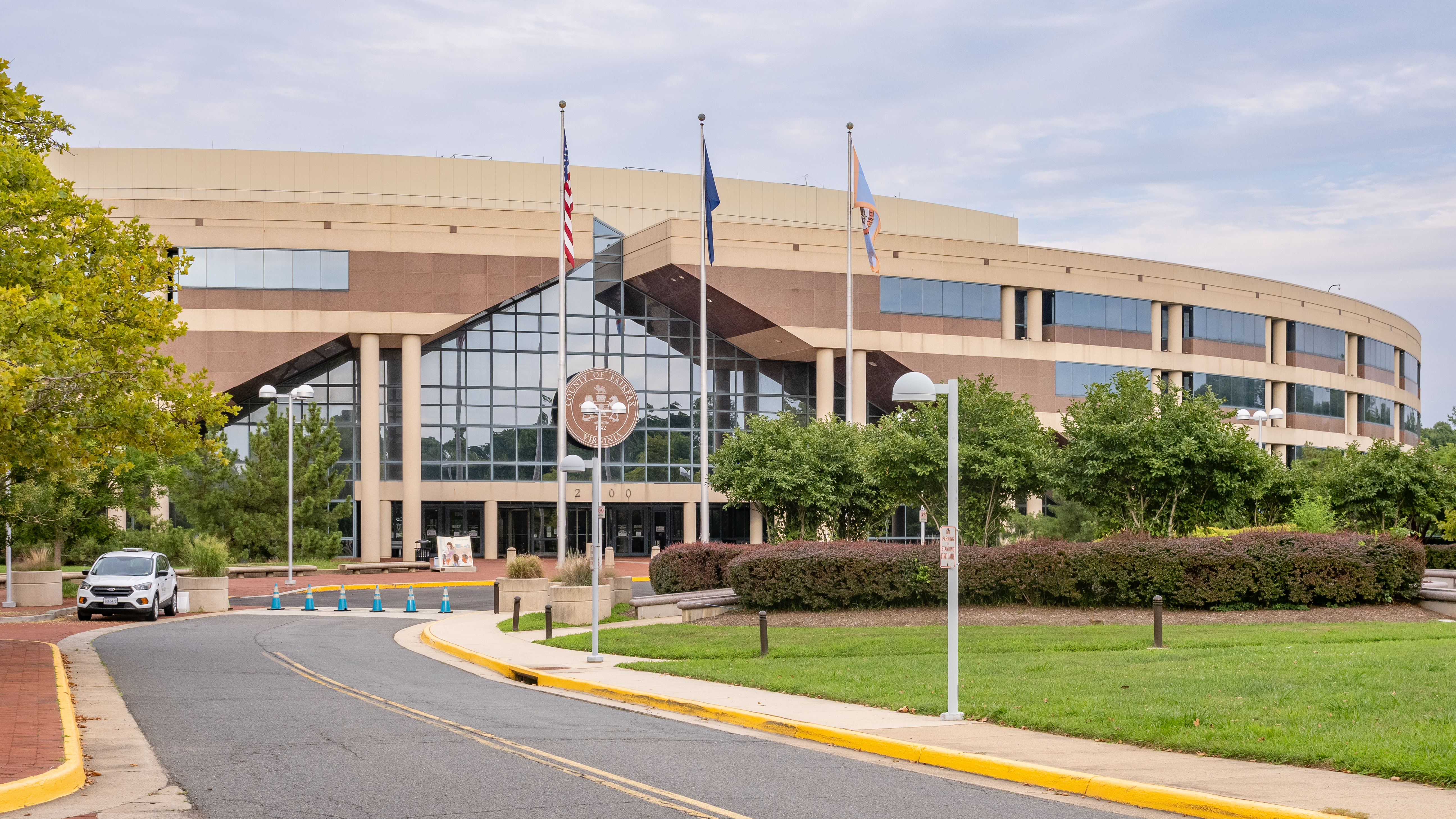 Large curved government building with flags on flagpoles and landscaped entrance driveway.