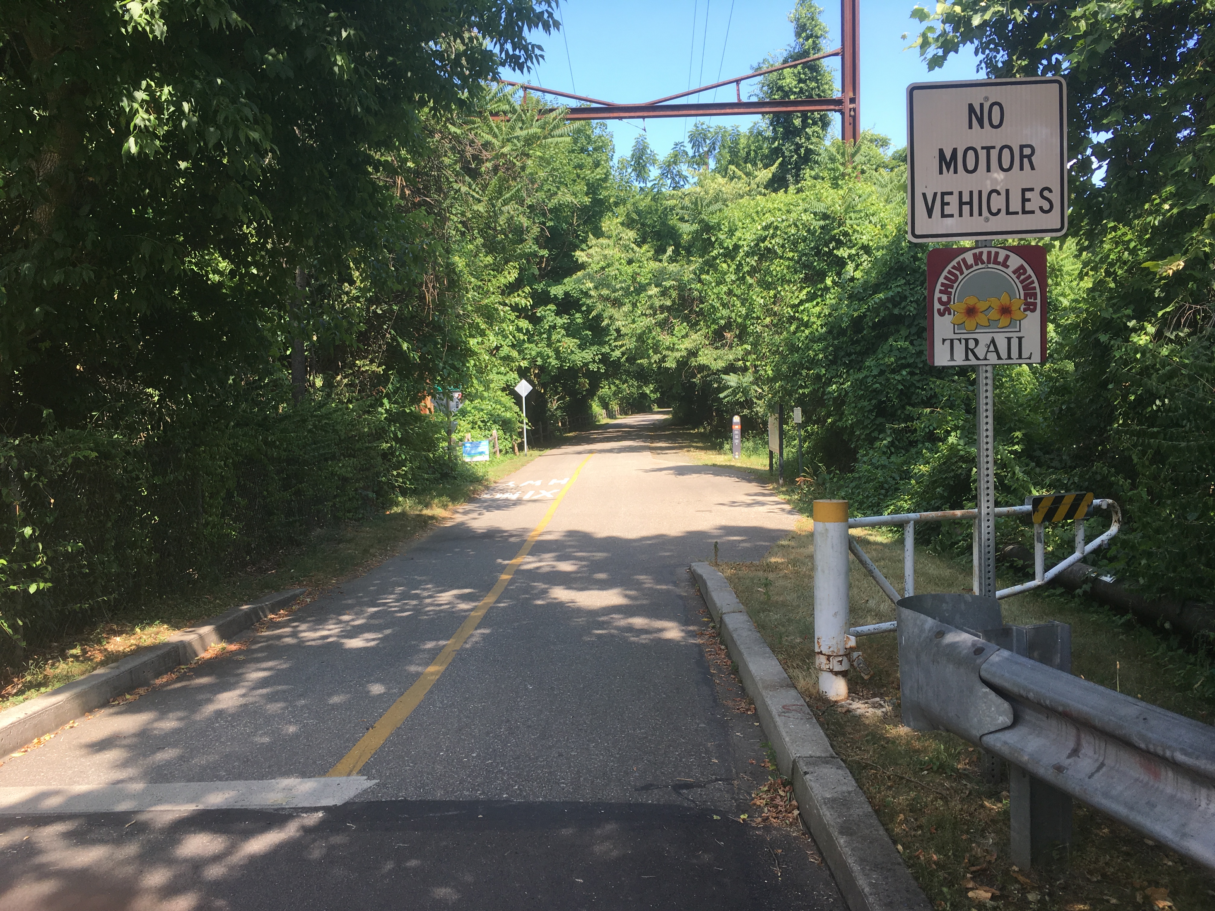 Paved multi-use trail with signage entering a tree-lined corridor.