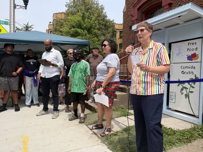 Woman in rainbow-striped shirt speaks into a microphone at an outdoor event beside a white pantry structure.