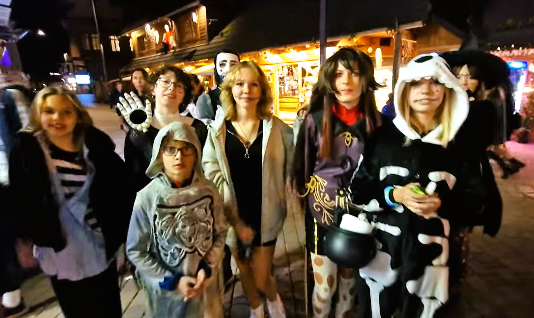 Group of people in Halloween costumes posing outdoors at night near illuminated storefronts.