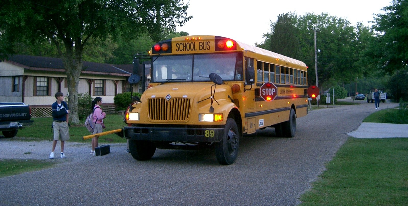 Two children with backpacks waiting beside a stopped yellow school bus on a residential street.