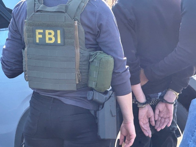 An FBI agent in a tactical vest stands next to a handcuffed person.