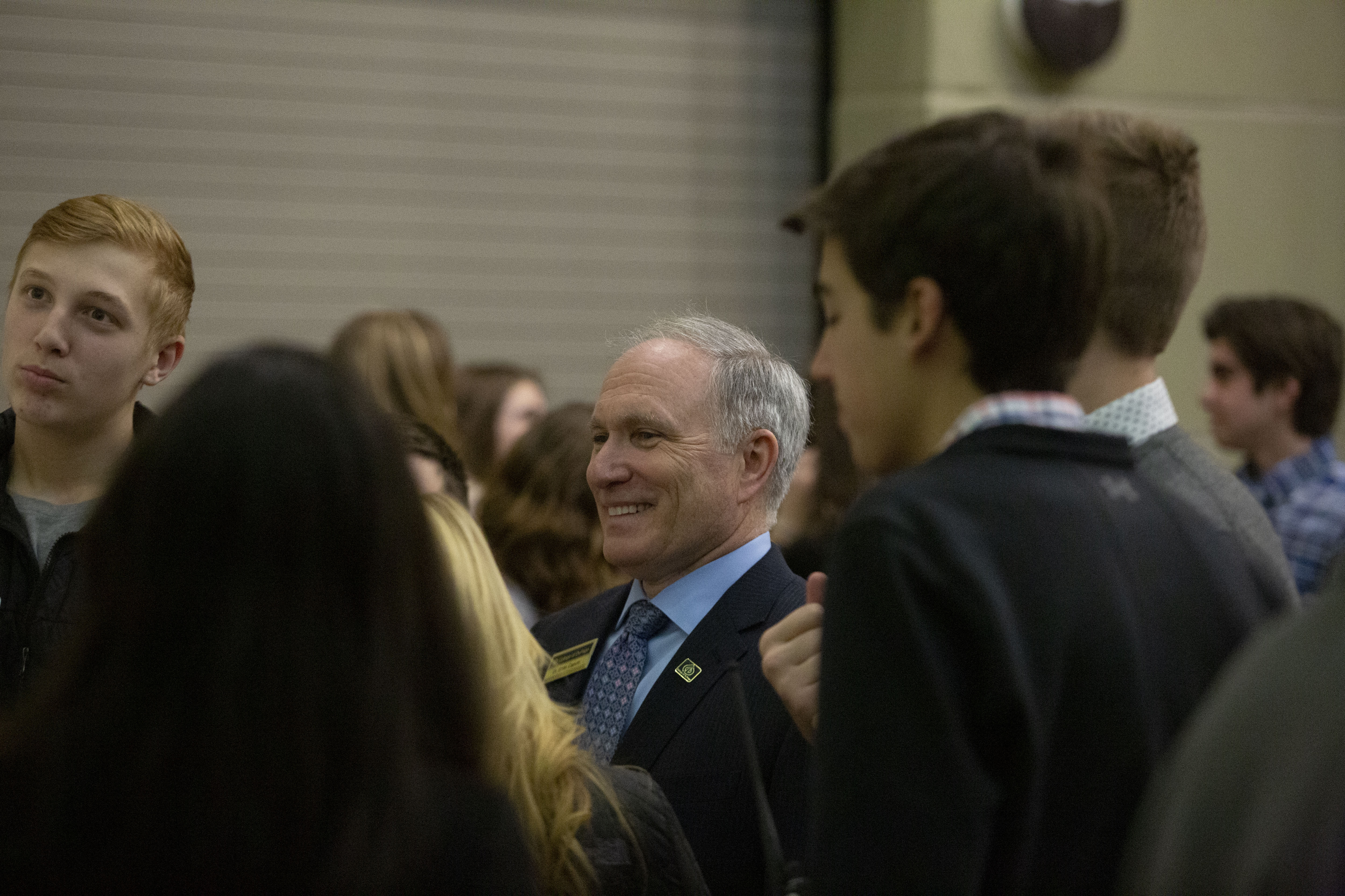 An adult in a suit smiles while talking with students at an indoor school event.