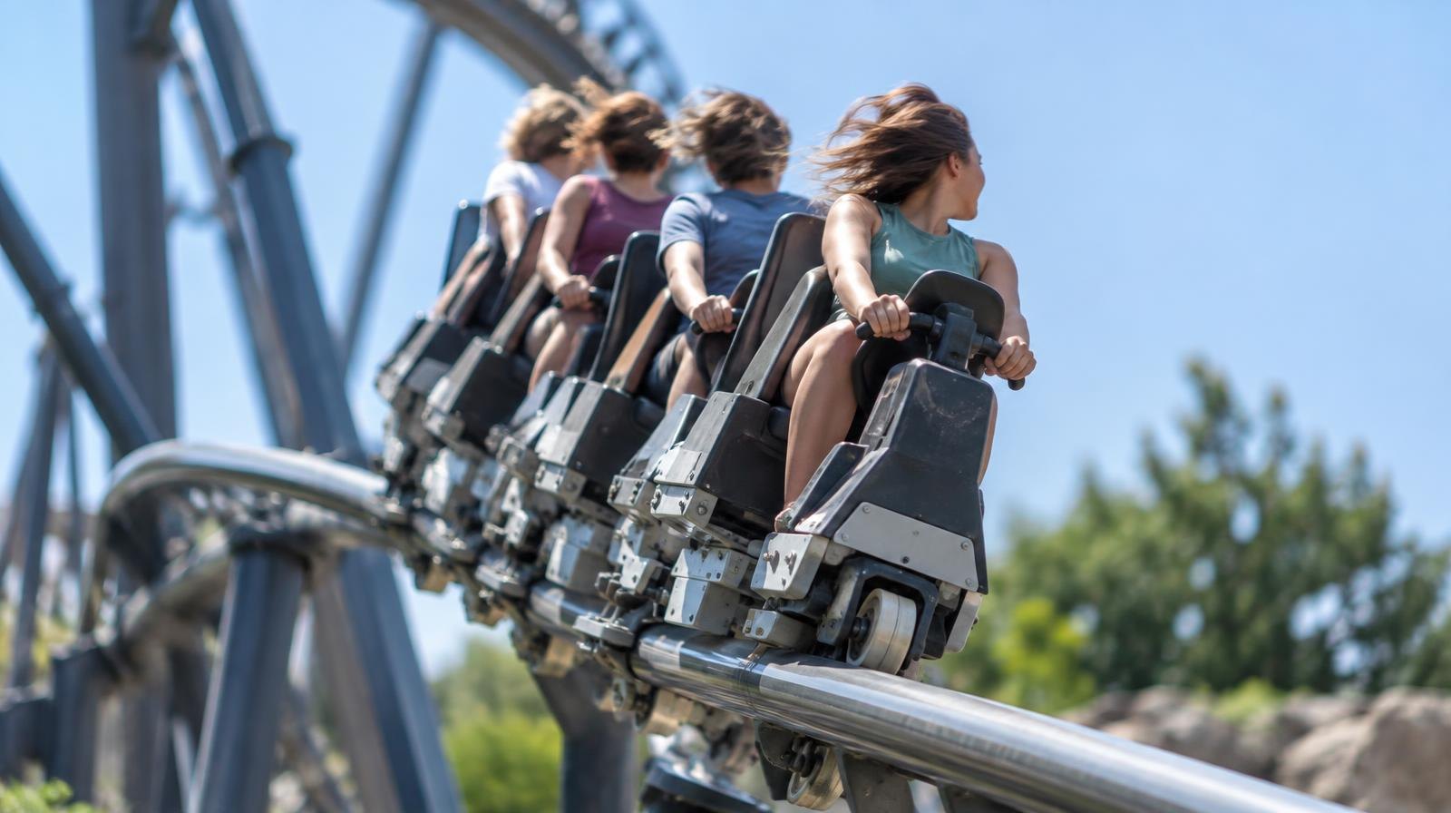 Roller coaster train with riders seated single-file above a narrow steel rail on a banked curve.