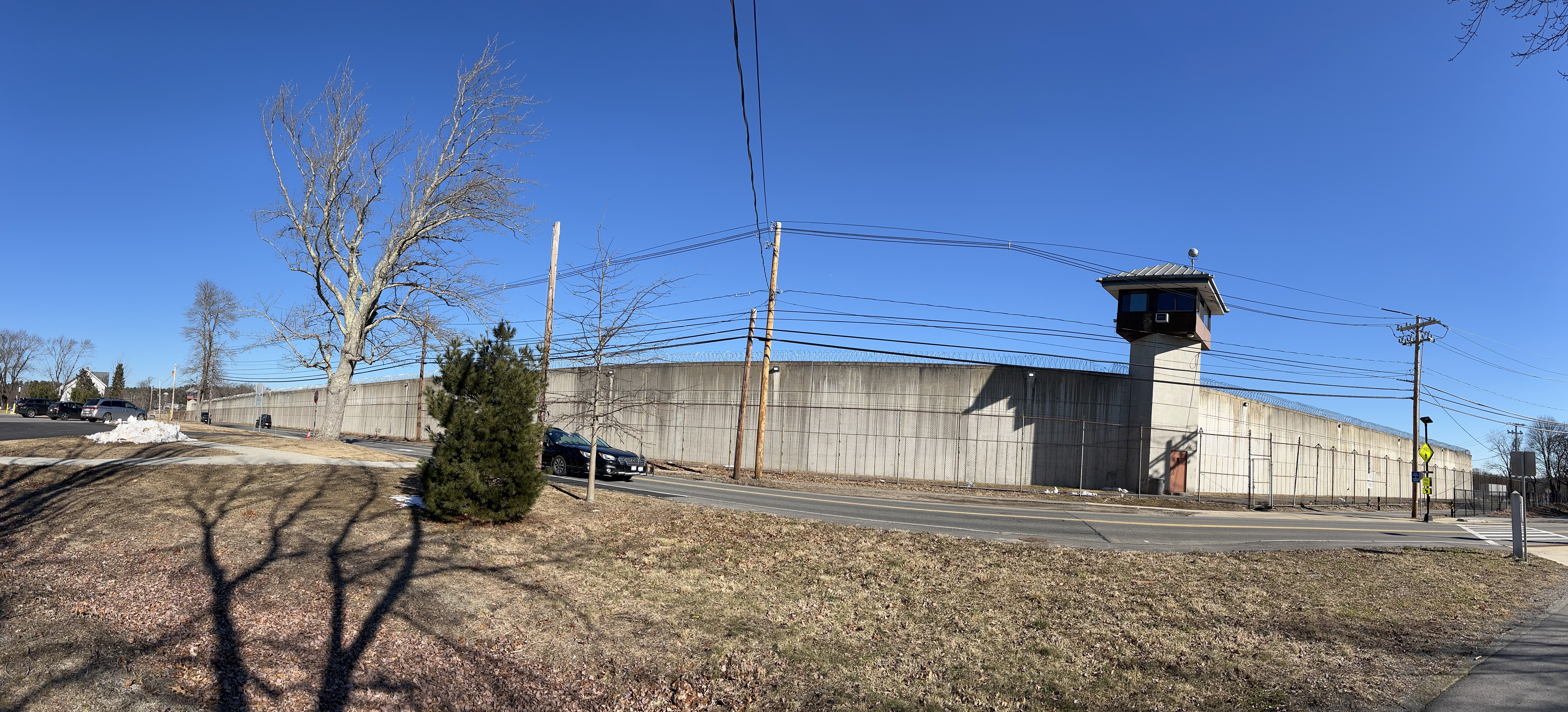 Tall concrete prison wall with guard tower alongside a road in winter.